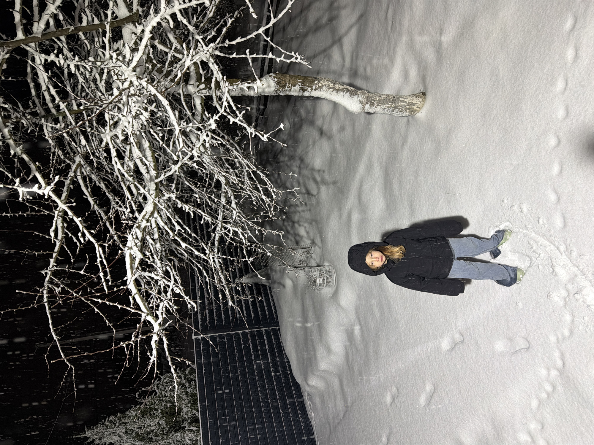 A girl is lying in the snow in what appears to be a backyard or garden at night. The area is covered in a thick layer of snow. The girl is wearing a dark jacket, jeans, and shoes, and the hood of her jacket is pulled up. There are visible footprints in the snow around her. To the left is a tree with snow-covered branches. Behind the tree is a dark fence. The overall lighting suggests it is either nighttime or a very overcast day. There is no definitive information in the image to confirm the location is Markovo, Russia.