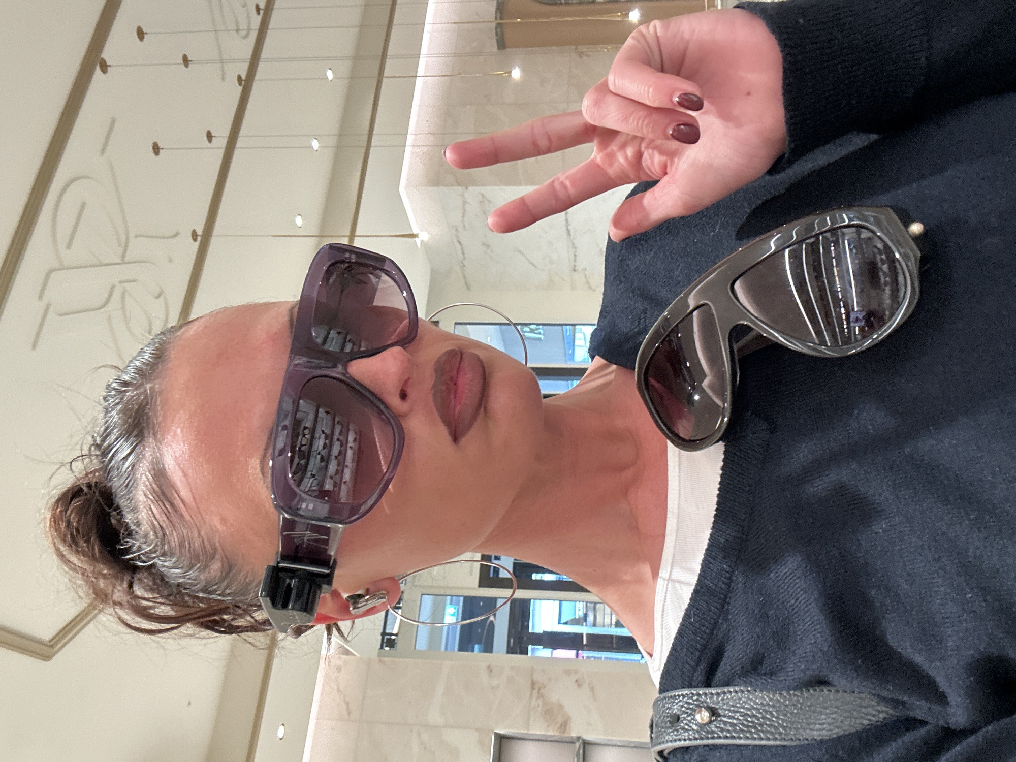 A woman indoors takes a selfie while wearing two pairs of sunglasses, one perched on her nose and the other on her black sweater. Her left hand is raised, fingers forming a peace sign. She wears large hoop earrings and has dark nail polish on her fingernails. The setting appears to be a retail environment, potentially an optician, as suggested by the reflection of sunglasses displayed in the lenses of the glasses she is wearing. The surroundings are modern, with a neutral color palette and linear design elements. The location is identifiable as London, United Kingdom, due to the reflection of a building exterior in her glasses, indicative of the city's distinctive architecture.