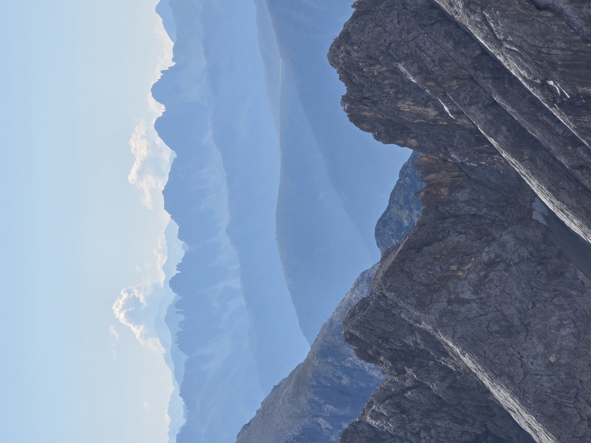 The image depicts a vast mountain range with multiple layers of peaks fading into the distance. The foreground is dominated by rugged, rocky mountain slopes, with sharp inclines and textured surfaces in shades of gray and brown. As the view recedes, the mountains become progressively bluer and softer, creating a sense of depth and atmospheric perspective. The sky is a pale blue with a few wispy clouds scattered across the upper portion. The scene is entirely natural, with no signs of human presence or structures. This image is likely a view of the Alps, possibly taken from a high vantage point in or around Garmisch-Partenkirchen, Germany, given the characteristic layered mountain landscape often seen in that region.