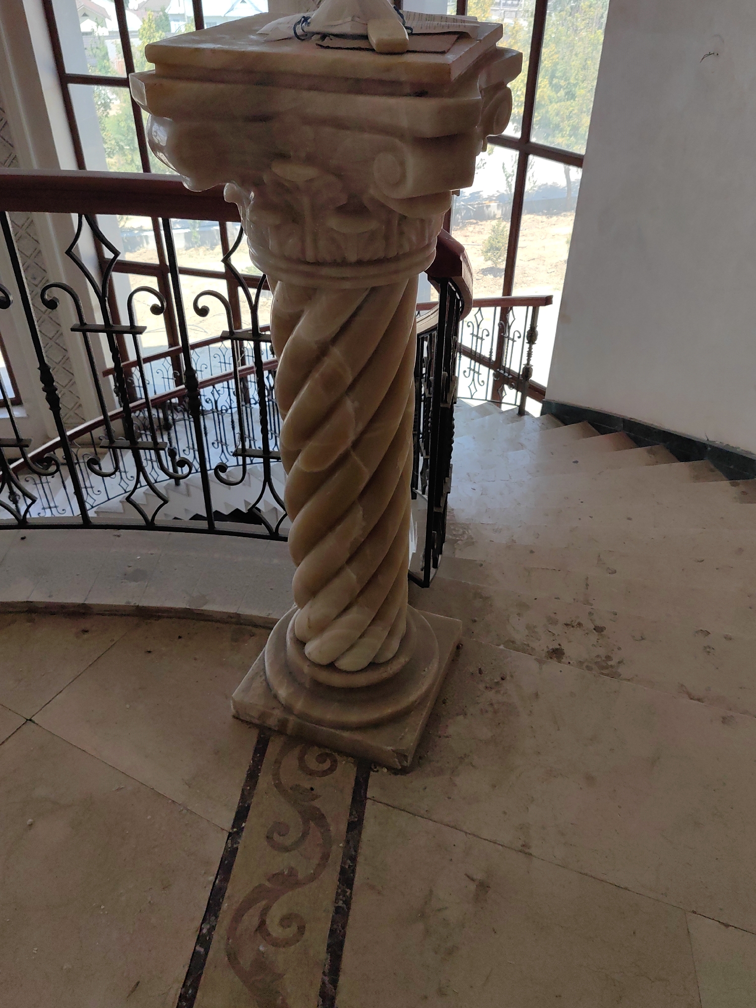 This image depicts an ornate marble column situated on a landing of a grand, spiral staircase. The column is carved with a spiral motif and topped with an elaborate capital. The staircase features a dark metal railing with decorative scrollwork. The flooring appears to be marble tiles with an inlaid decorative border. In the background, a large window reveals trees and some buildings, suggesting an indoor location with natural light. There are no people visible in the image. The scene is well-lit, with the marble column being the primary subject. The overall impression is of an elegant interior space, possibly a hotel, museum, or a large private residence. This specific location is not identifiable within the city of Shymkent, Kazakhstan, based on the provided visual information alone.