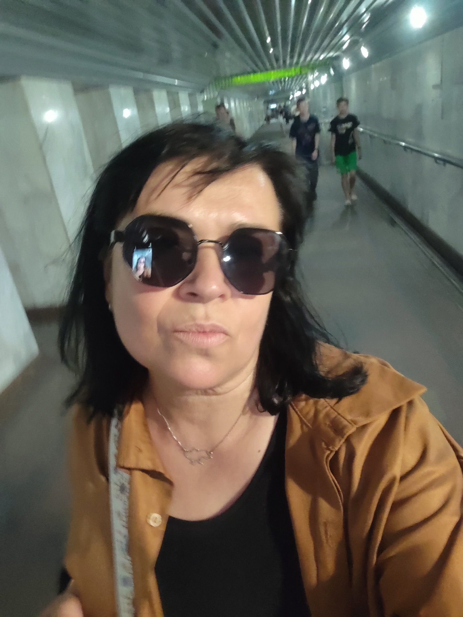 A woman wearing sunglasses and a brown jacket is taking a selfie in a tunnel. In the reflection of her sunglasses, a smaller image of herself is visible. The tunnel has a polished, reflective ceiling and walls, with lights spaced along the ceiling. Several people are walking in the background, some appearing as blurred figures. The woman has dark hair and is wearing a necklace.