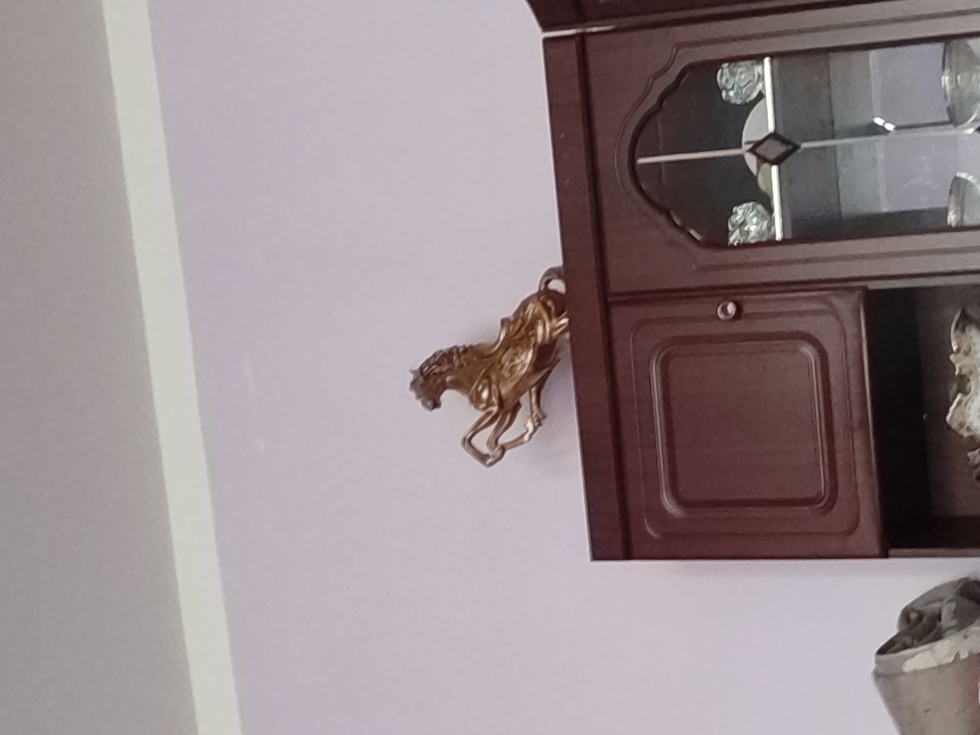 A golden statue of a rearing horse is positioned on top of a dark wooden cabinet. The cabinet has a glass door with decorative etching and appears to be part of a larger piece of furniture. The background is a pale pink wall. The image does not provide any information to suggest it is located in Baghdad, Uzbekistan.