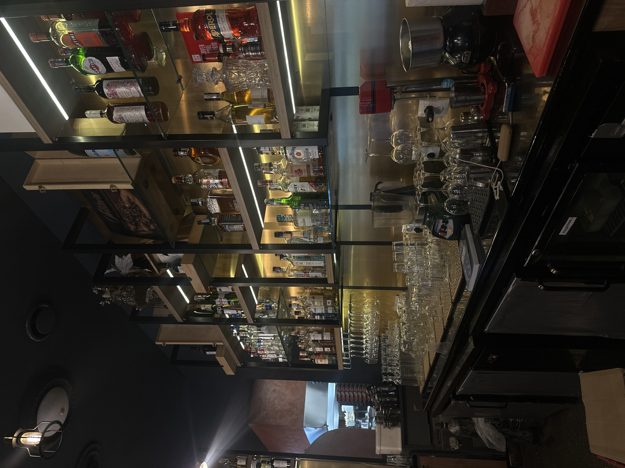 The image shows a dimly lit bar interior, likely in Sofia, Bulgaria, featuring a well-stocked liquor cabinet and a bar counter. The liquor cabinet, with multiple shelves illuminated by LED lights, displays a variety of bottled spirits and liqueurs, including brands like Martini, Aperol, Jack Daniel's, and Jim Beam. In front of the shelves, wine glasses are hung upside down. The bar counter is equipped with various items, such as a blender, a coffee grinder, and stacks of glassware. The overall ambiance is dark and moody, with a spotlight casting a beam across the scene from the upper left. No people are visible in the image.