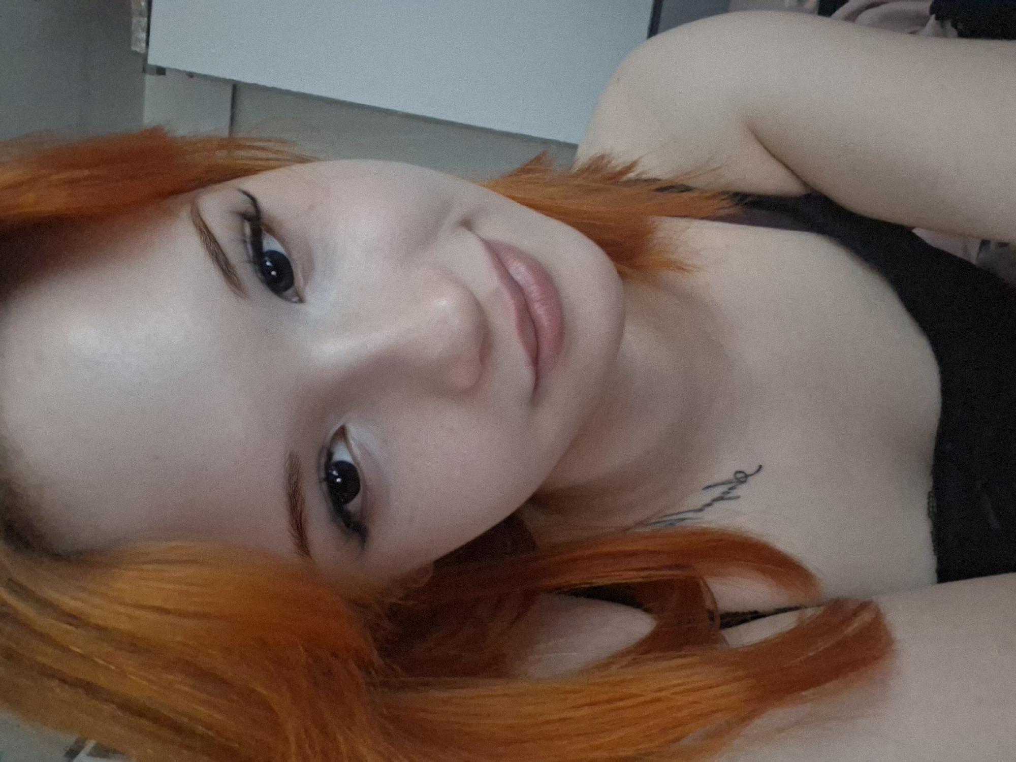 A close-up, angled portrait shows a fair-skinned woman with bright orange, shoulder-length hair lying down. Her head is tilted to the right, and she is looking directly at the camera with dark eyes. She has visible makeup, including eyeliner and lip gloss. She is wearing a black top. A tattoo is partially visible on her upper chest. The background is out of focus, showing a white or light-colored wall and a glimpse of bedding or clothing. There is no evidence in the image to indicate it was taken in the city of Chaikovsky, Russia.