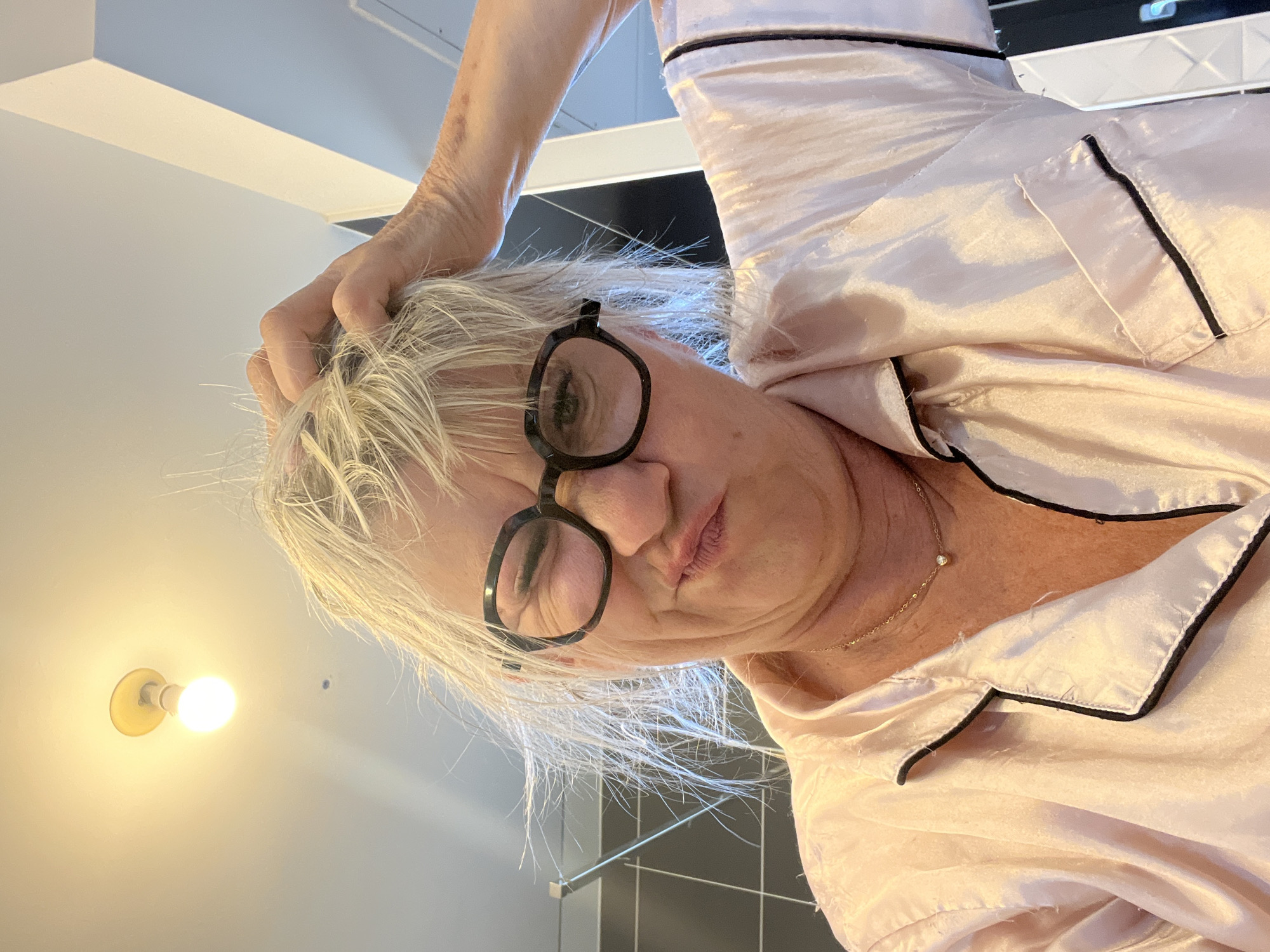 The image shows a woman with blond hair wearing glasses and a pink pajama top. She is looking at the camera and has her hand on her head, appearing to scratch it. The background shows a light fixture on the ceiling and a wall with black tiles. The image does not contain any information about the city of Chambœuf, France.