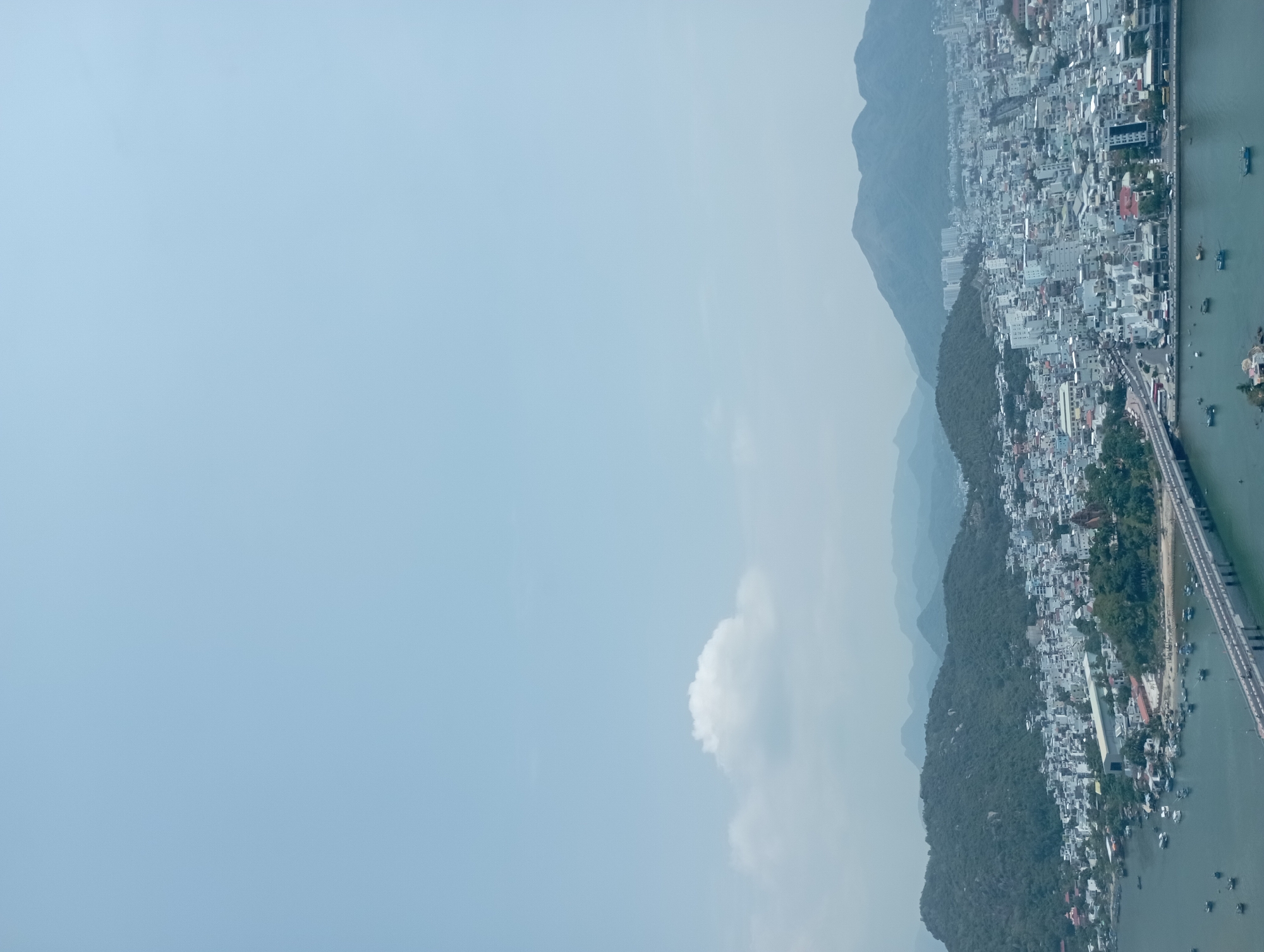 The image shows a coastal cityscape with mountains in the background, likely Nha Trang, Vietnam. The right side of the image contains a dense urban area with many buildings and a bridge extending across the water. Several boats are present in the water along the coast. Mountain ranges can be seen inland from the city. The left side of the image is primarily blue sky with a few clouds.