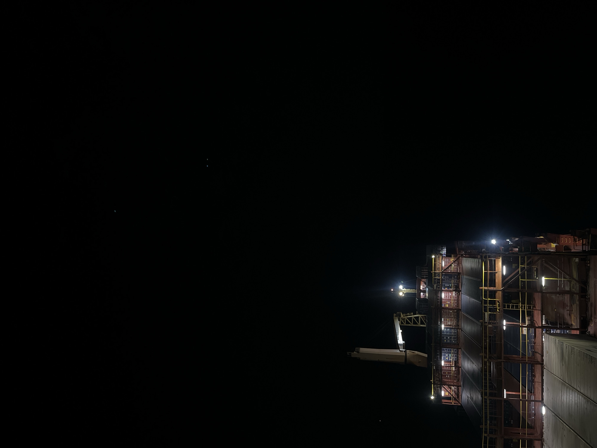 The image depicts a large structure, possibly a ship or building under construction, at night. The structure is illuminated by numerous lights, revealing a framework of scaffolding and containers. The sky is dark, with a few faint stars visible. A crane is attached to the structure. The location is described as Unknown, Unknown.