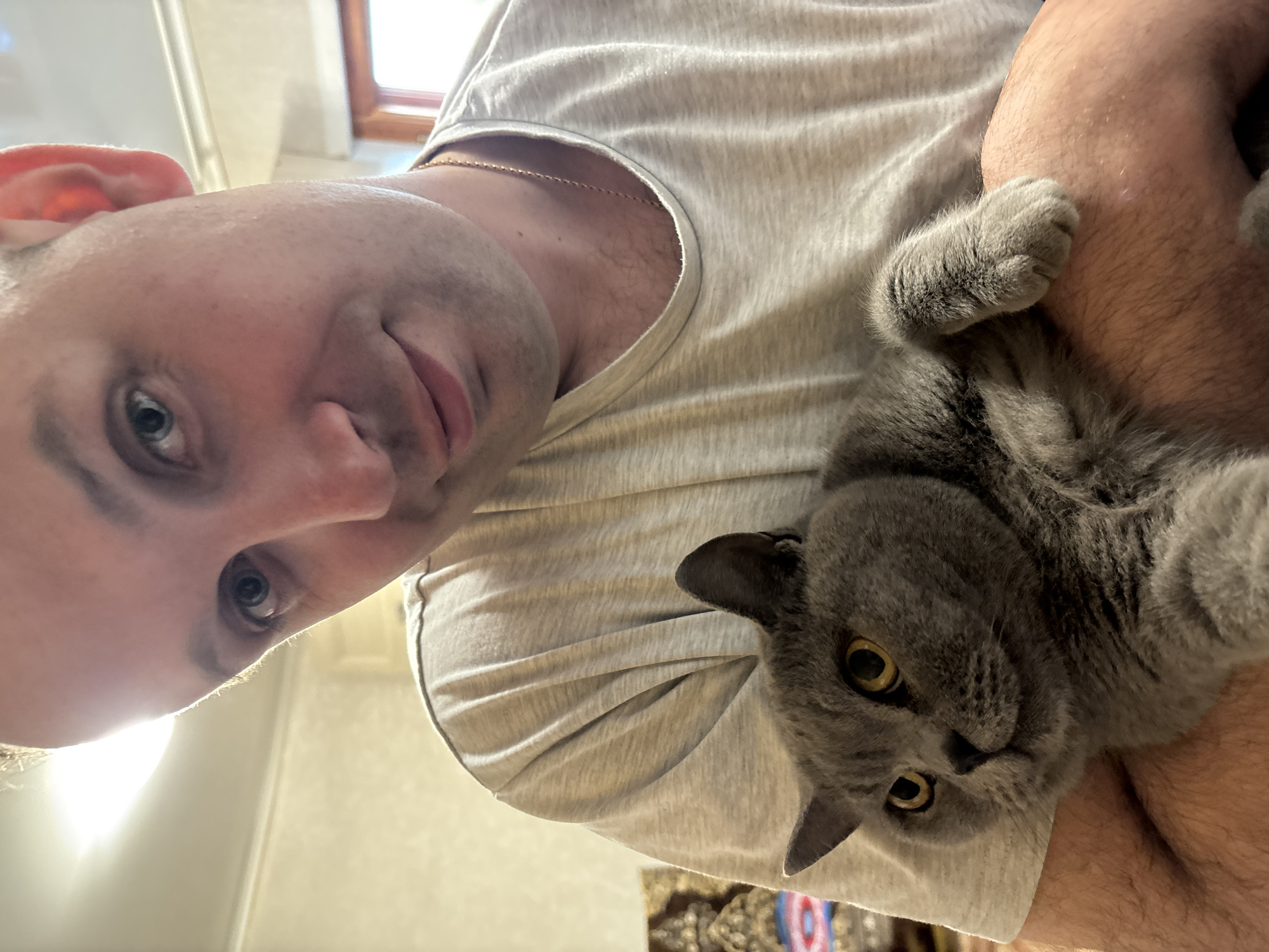 An indoor, close-up photograph taken in Rozhdestveno, Russia, features an adult male holding a grey cat. The man, with light skin, short light hair, and light-colored eyes, occupies the upper two-thirds of the frame, smiling faintly while looking directly at the camera. He wears a plain, light grey short-sleeved t-shirt and a thin gold chain around his neck. His lower right arm is visible, covered in light hair, supporting the cat. The cat, appearing to be a British Shorthair due to its dense blue-grey fur, round face, and prominent amber-yellow eyes, rests comfortably in the man's embrace. It looks slightly to the viewer's right with an alert expression, its front paws visible on the man's arm. The background is softly out of focus, revealing light-colored walls. A bright light source is positioned behind the man's head, creating a visible glow. Faint patterns of what appears to be a textile or wall hanging are visible on the far left, and a wooden window frame is partially visible on the far right.