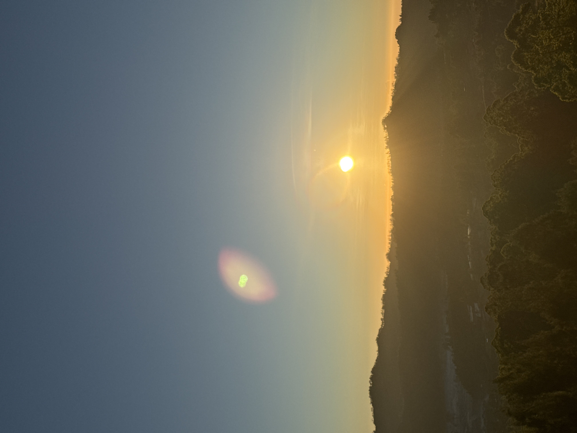 This image captures a sunrise over a mountainous landscape in Kintamani, Indonesia. The sun, a bright orb, is low on the horizon, casting a warm glow that transitions from deep orange near the sun to a pale yellow and then a clear blue at the top of the frame. The foreground is dominated by the silhouettes of lush green trees, with the layered shapes of hills stretching into the distance. A faint haze or mist softens the details of the further hills, adding depth to the scene. There are two distinct lens flares present: one a small, bright circular flare near the sun, and another larger, oval-shaped flare higher in the sky, exhibiting a pinkish hue with a greenish core. No people or specific structures are clearly discernible. The overall impression is one of a tranquil natural environment at dawn.