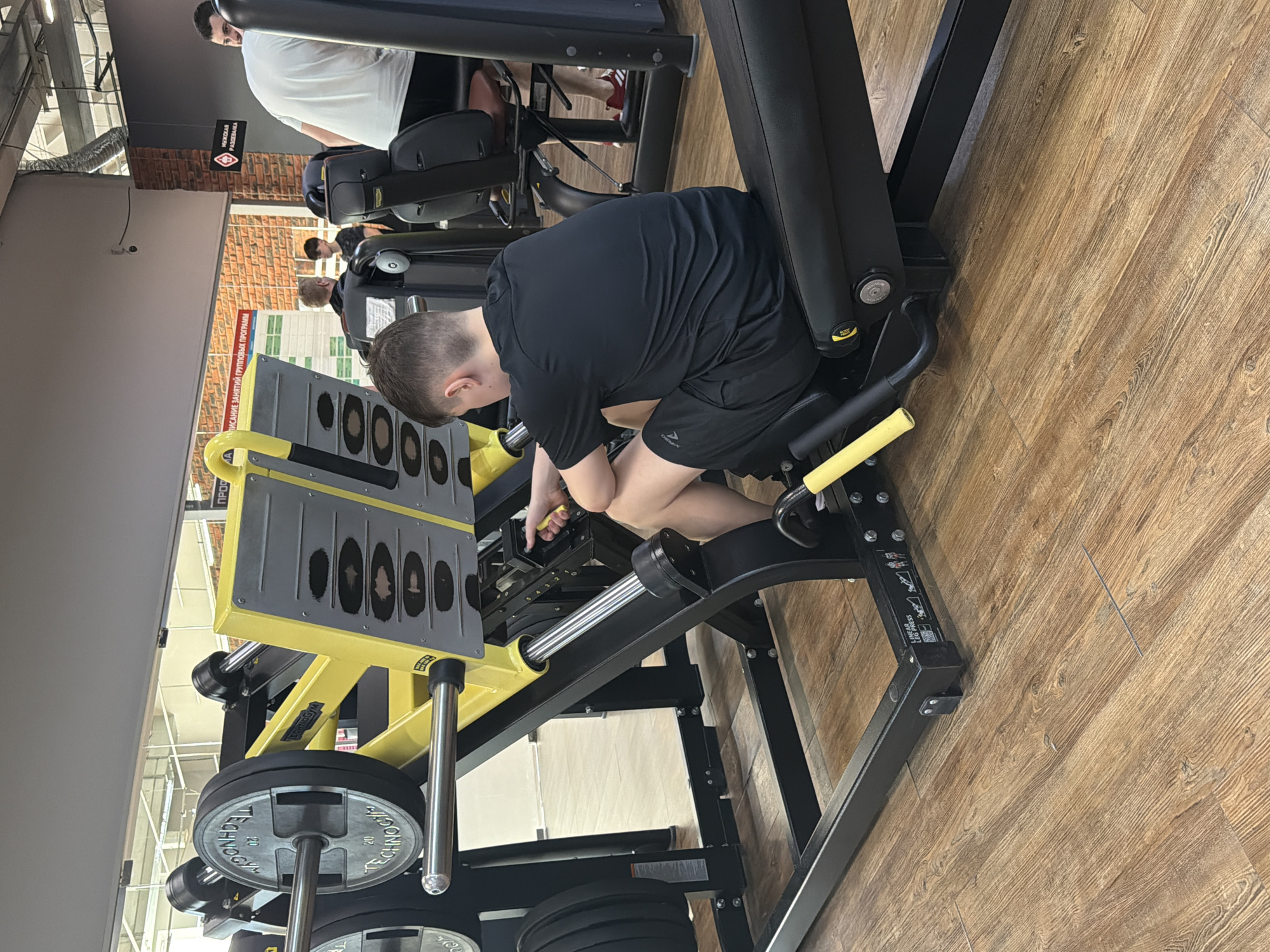 A person is using a leg press machine in what appears to be a gymnasium, possibly in Chaikovsky, Russia. The person is wearing a black T-shirt and black shorts. The leg press machine is black and yellow with weight plates loaded onto it. The floor is a light brown wood-grain. Other people are present in the background.