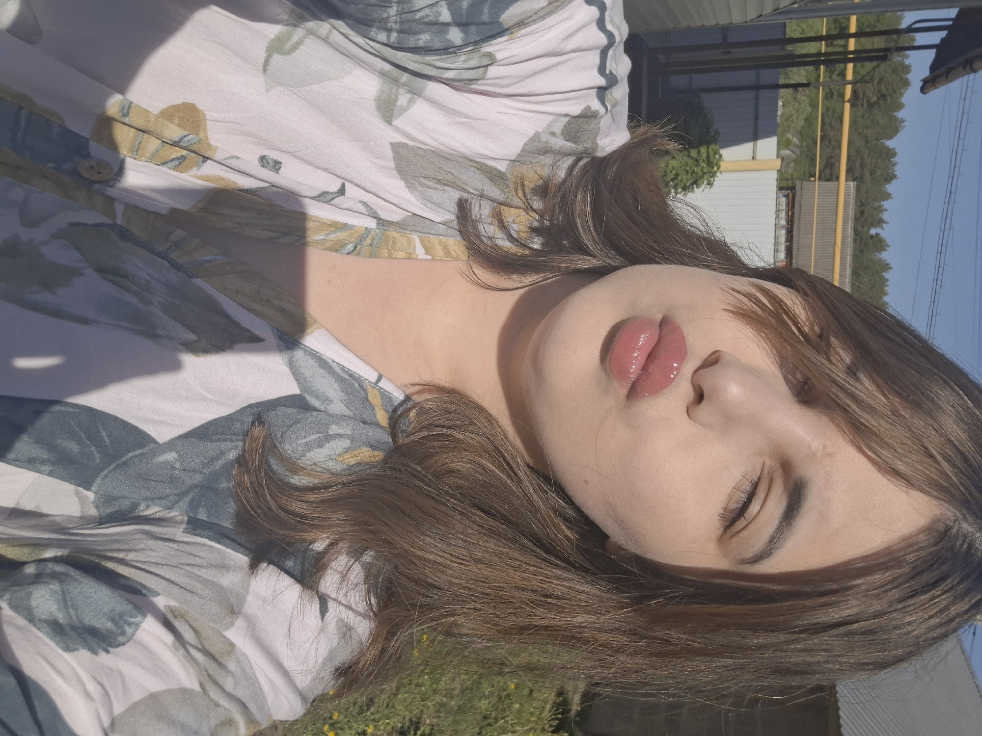 The image depicts a young woman with light skin, light brown hair, and full lips posed in an outdoor setting. She is wearing a floral-patterned garment. The background shows a metal fence, a building with a corrugated metal roof, and trees. The location appears to be a sunny outdoor area in what may be a residential or semi-urban environment. The information about the specific location in the city of Амзя, Russia is assumed based on available data.