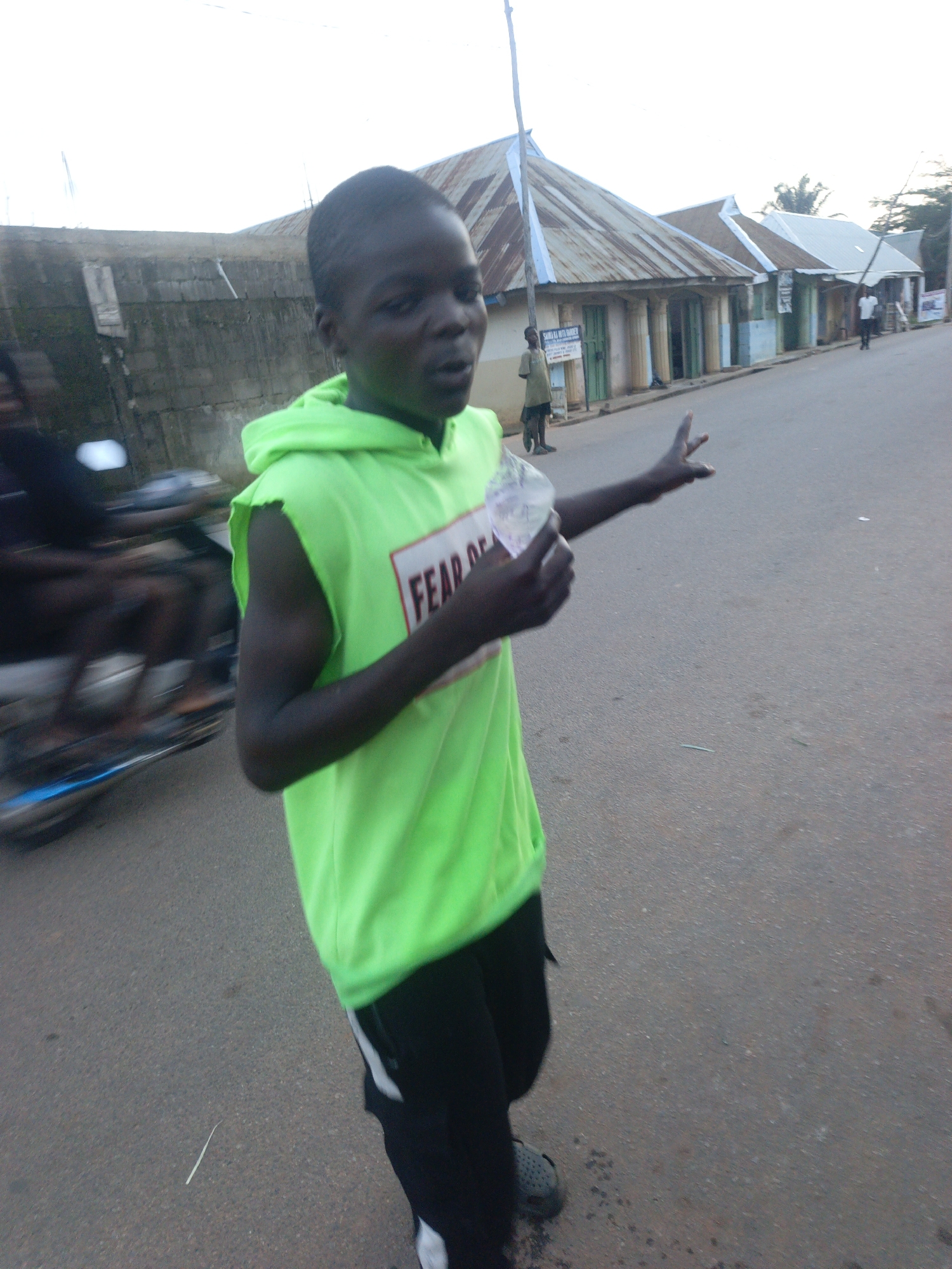 A mid-shot captures a young person with dark skin standing on an asphalt road in Akwanga, Nigeria. They wear a bright green hooded sleeveless shirt with the words "FEAR OF" partially visible on the front, and black shorts with a white stripe down the side. They also wear gray shoes. The person is holding something transparent in their left hand, and their right arm is extended. A motorcycle with two occupants is partially visible on the left. Behind them is a building with a stone wall. In the background, along the right side of the image, are single-story buildings with corrugated metal roofs and storefronts. There are people in the background walking along the street. The sky is overcast.