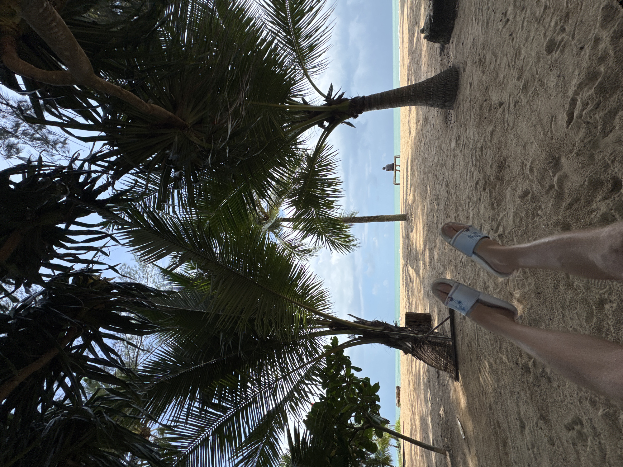 The image depicts a tropical beach scene in Ban Khuk Khak, Thailand. The foreground shows a person's legs and feet, wearing light-colored slides, resting on the sand. The perspective is from the ground looking up and out towards the ocean. Several palm trees with broad fronds dominate the midground, casting dappled shadows on the sandy beach. In the distance, a lone individual sits on a wooden bench facing the turquoise ocean and a slightly overcast sky. The environment suggests a relaxed, seaside atmosphere.