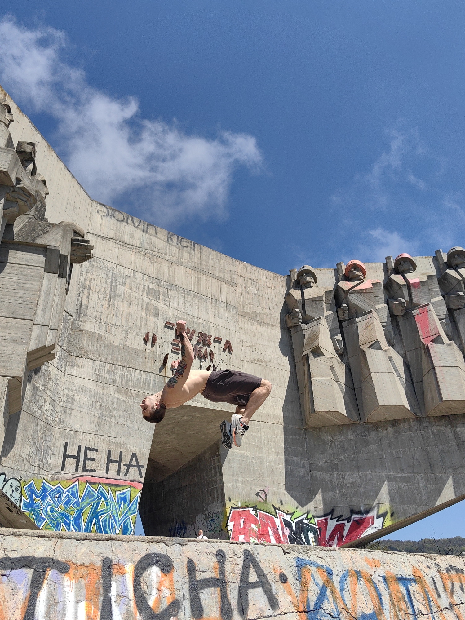 A man is performing a backflip in front of the Buzludzha Monument in Varna, Bulgaria. He is shirtless, wearing shorts and sneakers, and has tattoos on his arm. The monument is a large concrete structure with graffiti and Soviet-era sculptures. The sky is blue with some clouds.