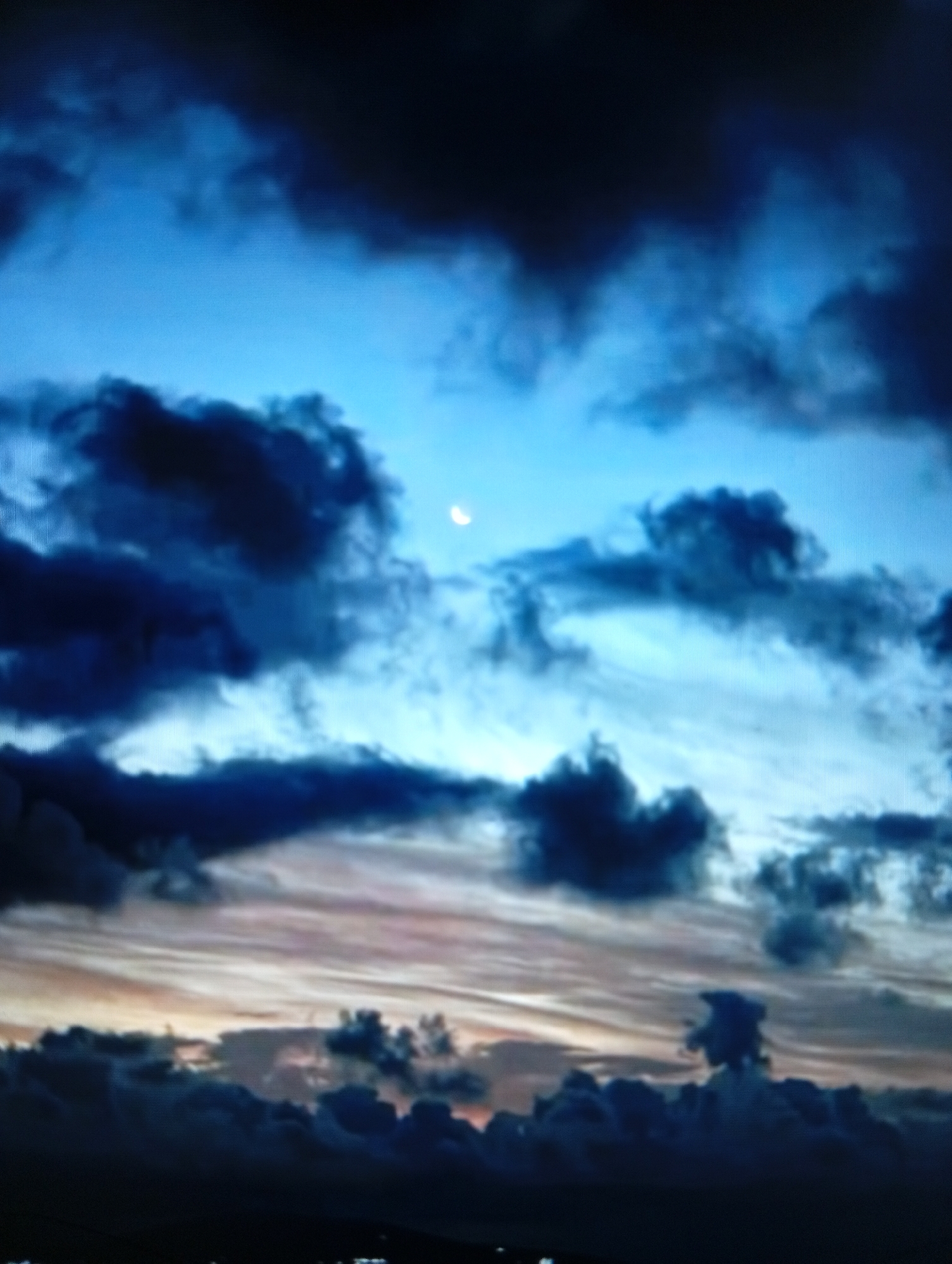 A low-angle, vertical shot captures a dramatic twilight sky filled with dark, turbulent clouds. A sliver of a crescent moon is visible in the upper portion of the frame against a bright blue sky. The lower part of the image shows a gradient of colors from dark blue to orange and yellow, suggesting the last light of sunset. The clouds in the foreground are dense and dark, obscuring any view of the ground. The scene depicts a stormy or overcast evening sky. There is no discernible location in the city of Esik, Kazakhstan, or any people, subjects, or objects within the frame. The environment depicted is purely atmospheric.