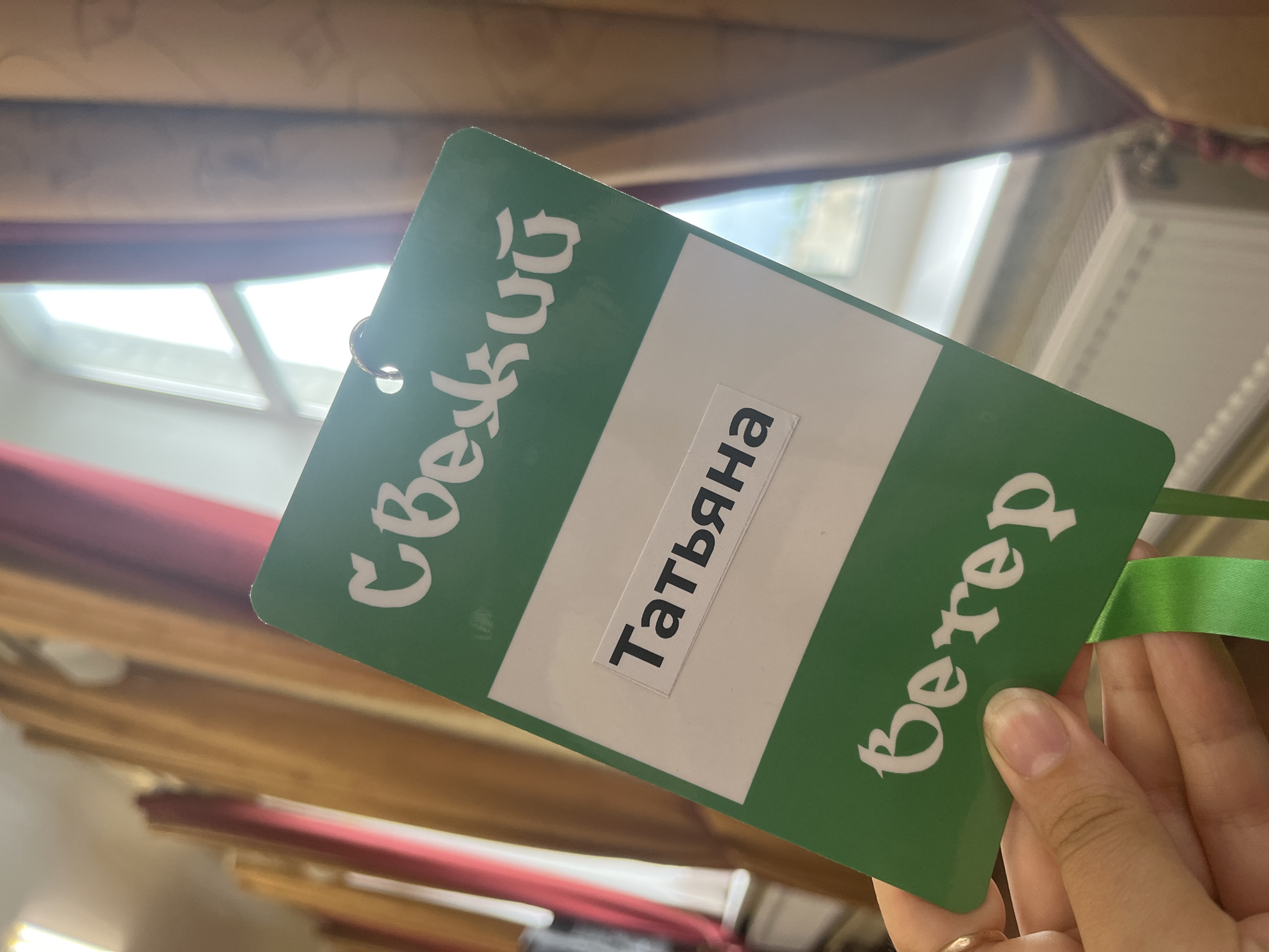 A person is holding up a green identification badge that reads "Свежий" on the left side, "Татьяна" in the middle, and "Вечер" on the right side. The text is written in white, and the badge has a green ribbon attached to it. The background is blurry, but it appears to be a room with light-colored walls and a window. The location is Unknown, Unknown.
