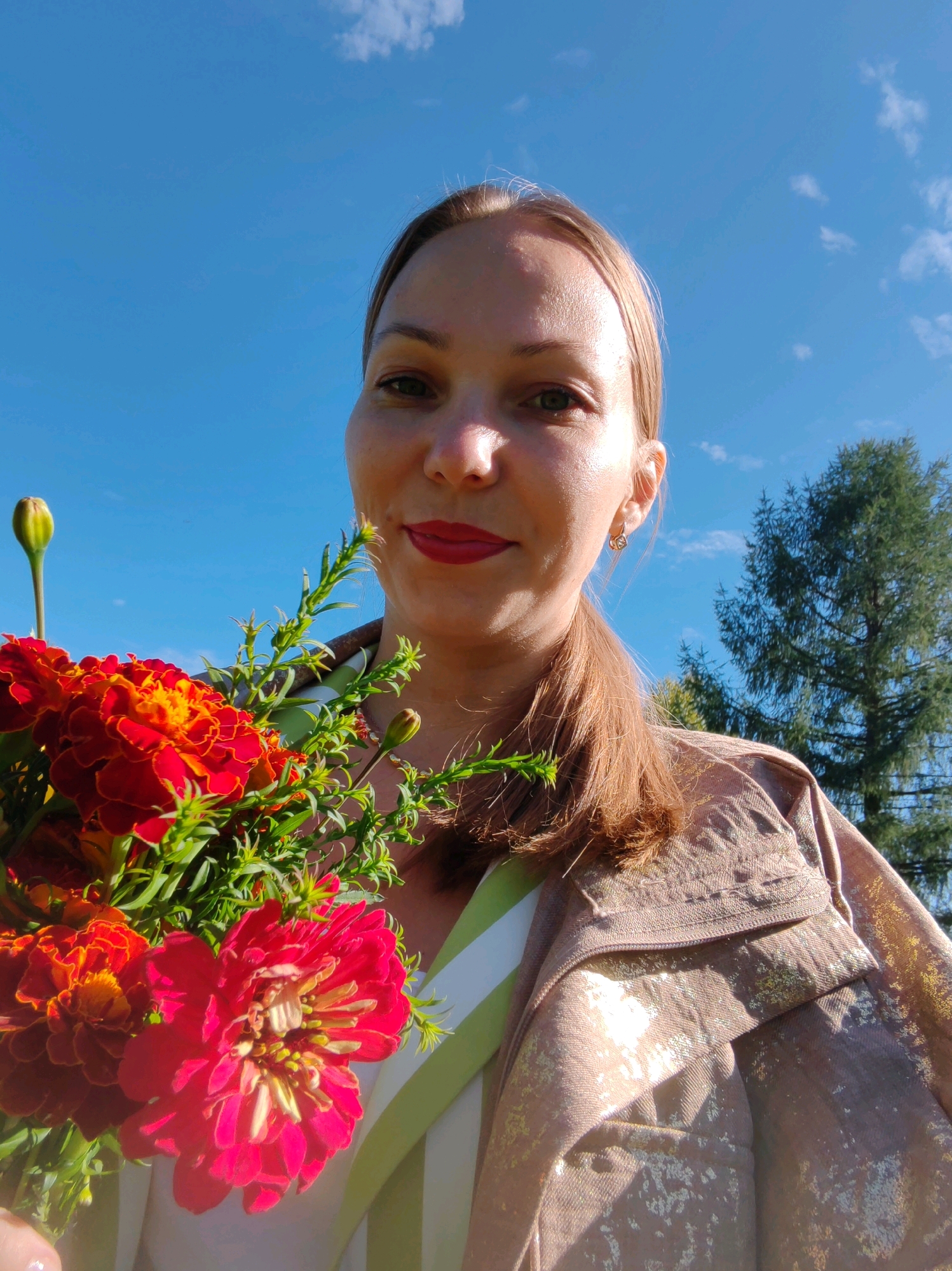 A woman with light brown hair, wearing a light brown jacket with gold flecks, holds a bouquet of marigolds. She is smiling at the camera, with red lipstick on her lips. The background shows a bright blue sky with a few clouds and a tall evergreen tree. The image appears to be a selfie taken outdoors. No specific location in the city of Чухлэм, Russia is identifiable from the image.