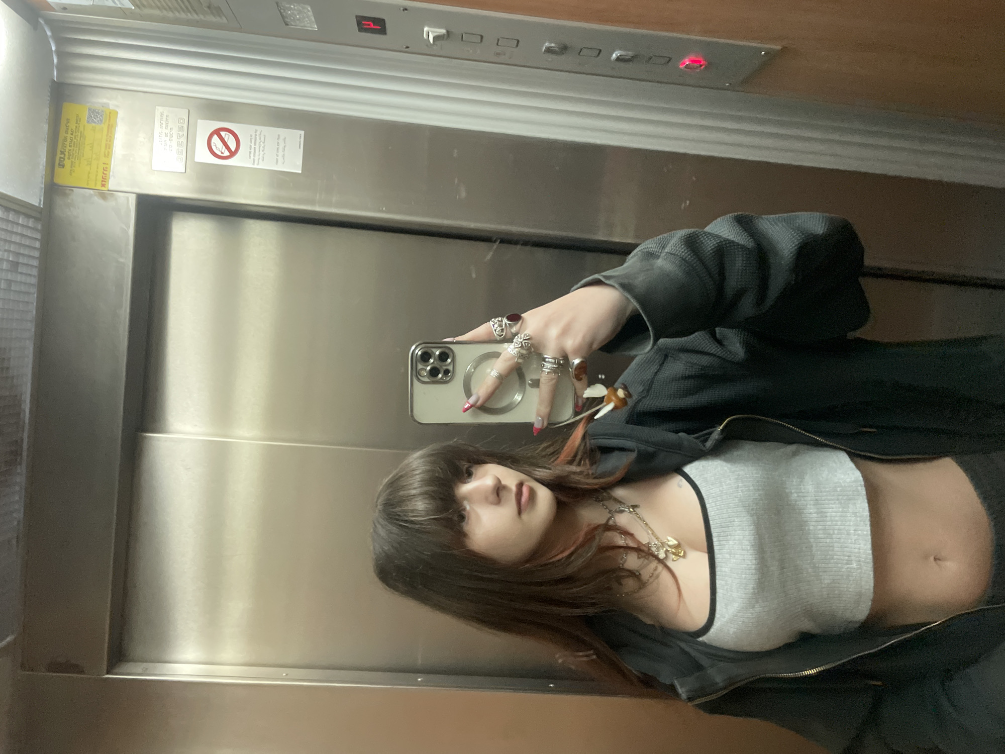 The image displays a young female inside an elevator, capturing her reflection in a mirror selfie. She possesses long brown hair with bangs and subtle reddish highlights, dressed in a grey ribbed crop top and an open dark waffle-knit hoodie. Multiple rings adorn her fingers, and several layered necklaces with various pendants are visible, including a prominent gold-colored charm. She holds a silver smartphone, equipped with a multi-lens camera array and a small decorative charm, positioned to photograph her reflection.
The elevator interior features brushed stainless steel doors and a left wall, contrasted by a light brown paneled wall on the right. An elevator control panel on the right wall displays a red illuminated "H" (with "P" below it) floor indicator and a red-lit button. Affixed to the door frame are a yellow warning sticker with Hebrew text, including "אמניף" (Amnif, an Israeli elevator company) and "חובה!" (Danger!), a metal plaque possibly reading "D.G. TECH", and a "No Smoking" symbol with additional Hebrew instructions. Based on the Hebrew signage and company identification, the location is a building in Petah Tikva, Israel.