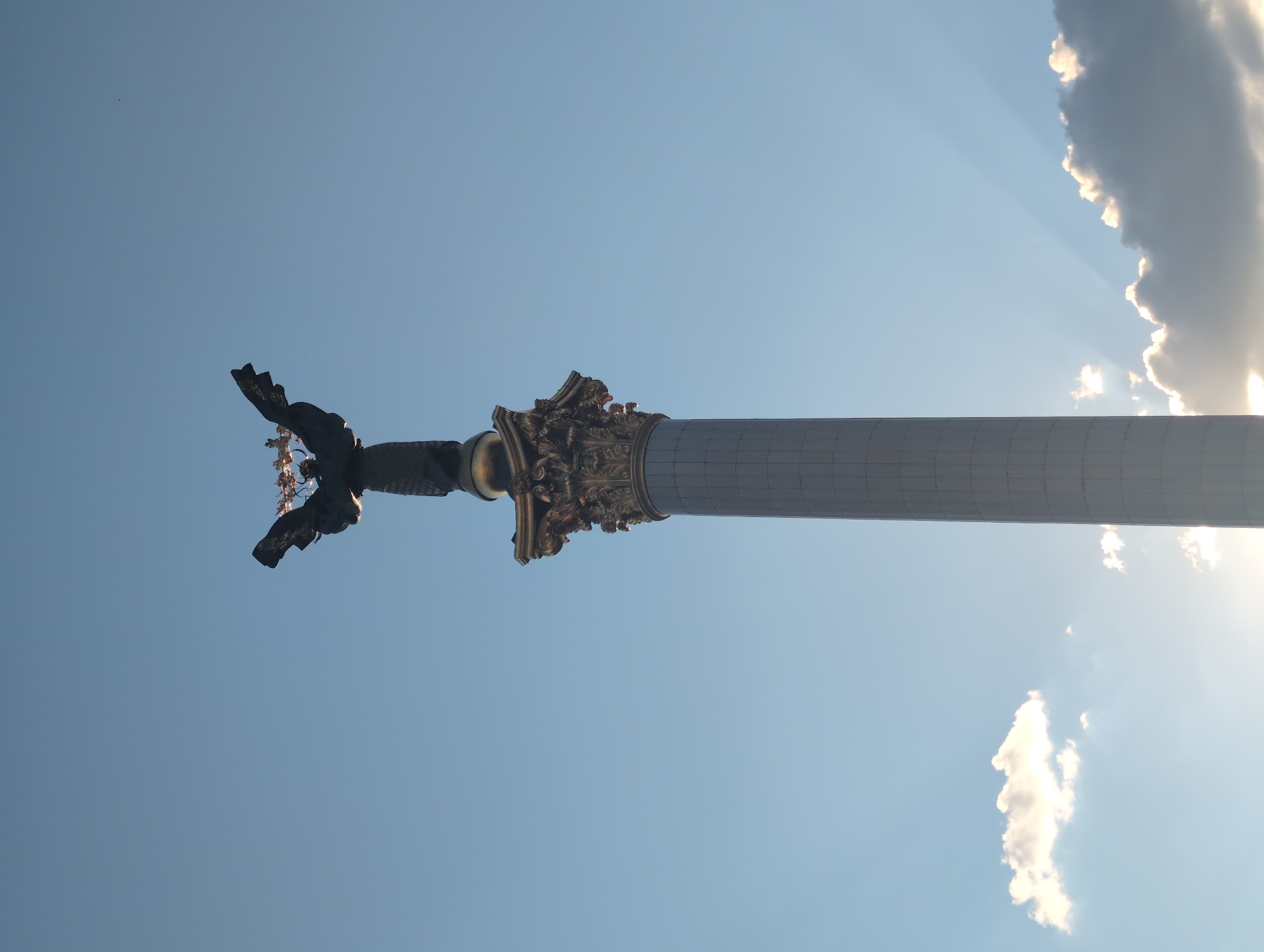 The image shows the Independence Monument in Kyiv, Ukraine, against a clear blue sky with scattered clouds. The monument is a tall, white, fluted column topped with a bronze statue of Berehynia, a Slavic goddess, holding a laurel branch. The statue is silhouetted against the sky, with the sun's rays visible behind the clouds on the right side of the image. The monument is located in Maidan Nezalezhnosti (Independence Square) in Kyiv.