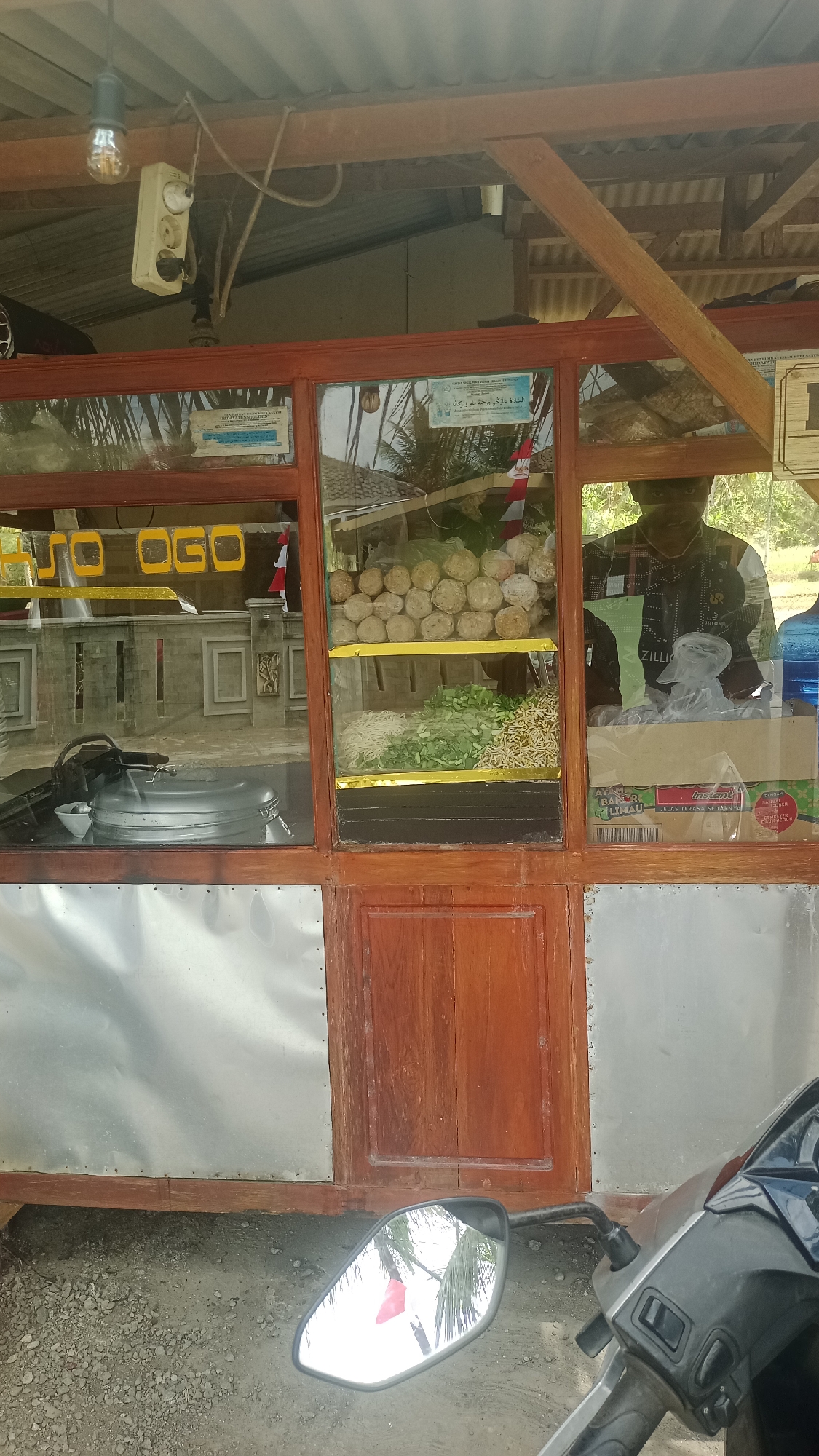 In the city of Cibeber, Indonesia, a food stall is depicted. The stall has a wooden frame and glass windows displaying its offerings. Prominently visible are stacks of meatballs, fresh vegetables, and noodles. A person wearing a dark shirt is partially visible through the glass, seemingly attending to the stall. The stall is equipped with a large metal pot, likely used for cooking. The exterior of the stall is covered with corrugated metal panels. In the foreground, a motorcycle's mirror reflects the surrounding environment, including palm trees and the sky. The ground is covered with gravel.