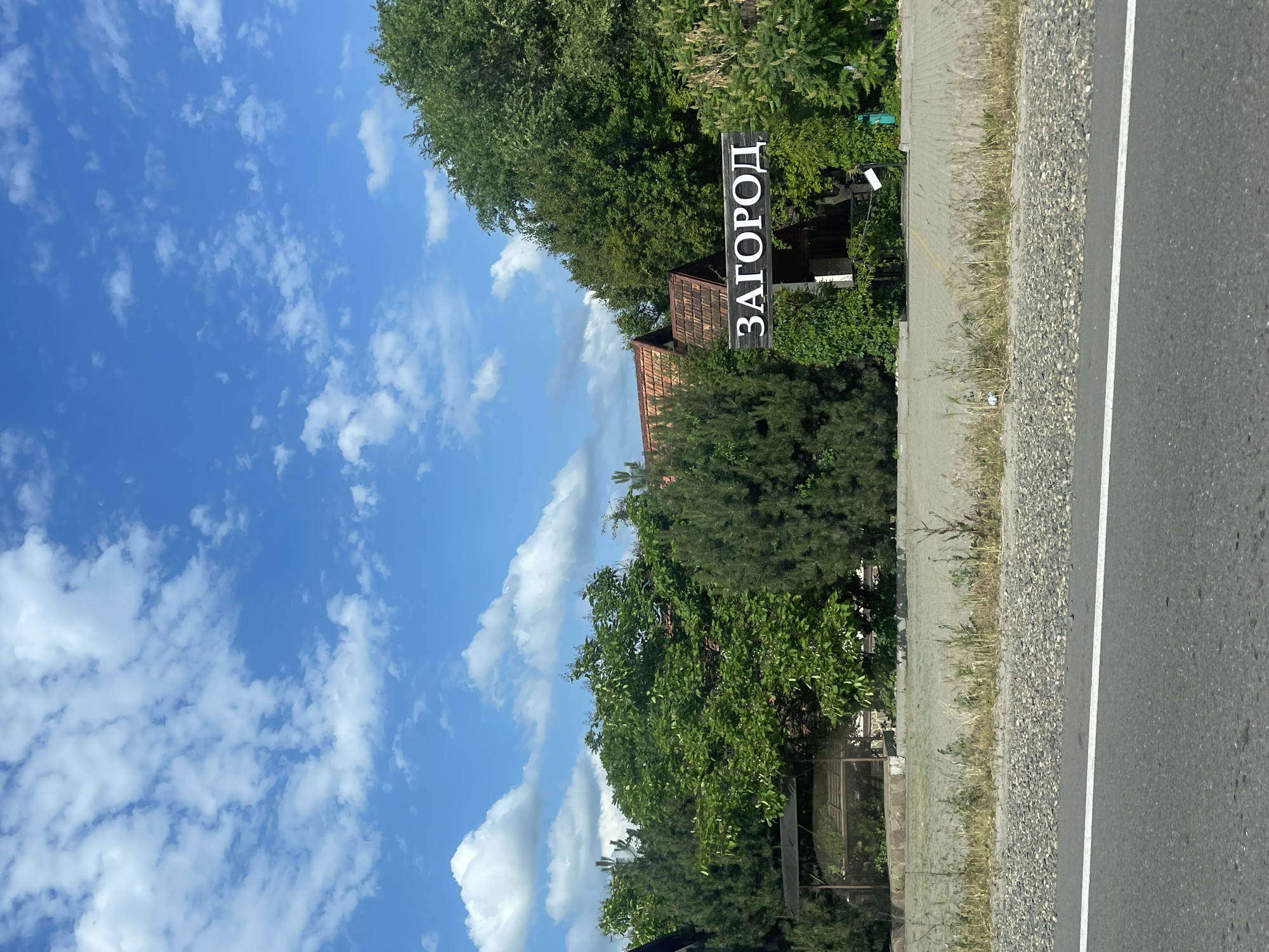 A vertical image shows a street scene in Donskoe, Russia, on a sunny day with a bright blue sky and scattered white clouds. On the right side of the image is a paved road with a white line marking the edge. Adjacent to the road is a gravel shoulder, followed by a paved pedestrian walkway or sidewalk. Beyond the walkway, there is lush green foliage and trees, interspersed with a few buildings with terracotta roofs.
A notable object in the scene is a dark wooden sign with white Cyrillic text that reads "ЗАГОРОД," meaning "out of town" or "suburb." The sign is placed amongst the trees, indicating the edge of a built-up area or perhaps an entrance to a residential community. The overall atmosphere is peaceful and suggests a rural or semi-rural setting. The lighting indicates it is daytime, likely mid-morning to late afternoon. There are no people visible in the image, and no specific activity is taking place. The focus is on the natural elements of trees and sky, combined with the man-made elements of the road and the directional sign.