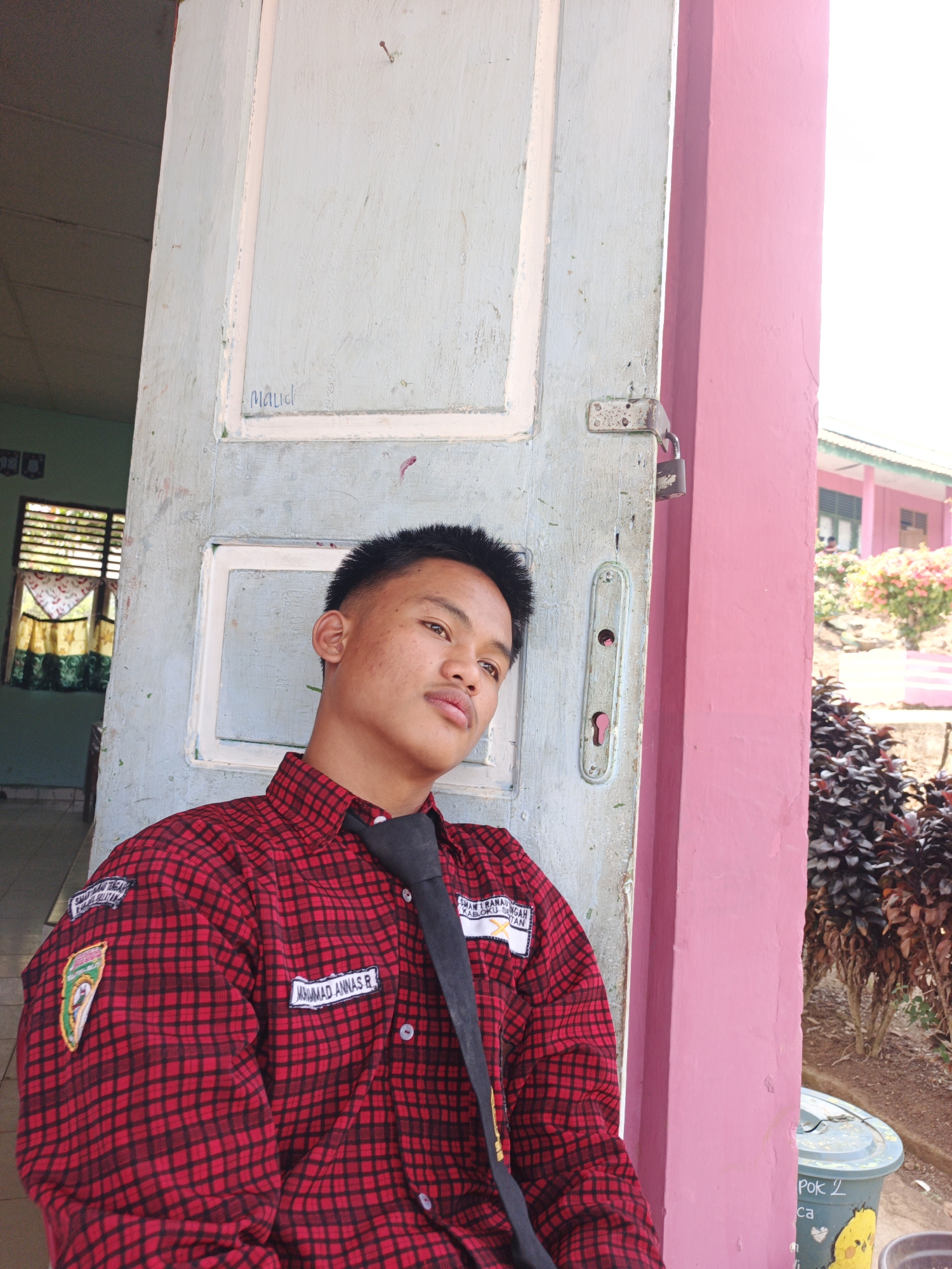 In Tanjung Setia, Indonesia, a young male individual is captured in a medium shot, leaning against a light blue door with white trim. He is wearing a red and black plaid collared shirt with a black tie. His hair is short and styled upwards, and he has a slight stubble. The door, weathered and slightly peeling, has a metal lock and latch attached. To the right of the door, a bright pink pillar stands out. In the background, beyond the pillar, a building with pink and green accents is visible, along with lush green foliage and trees. Sunlight casts shadows, indicating it is daytime. The overall scene suggests a casual, outdoor setting, possibly near a school or residential area, given the uniform-like attire of the subject and the presence of the building.