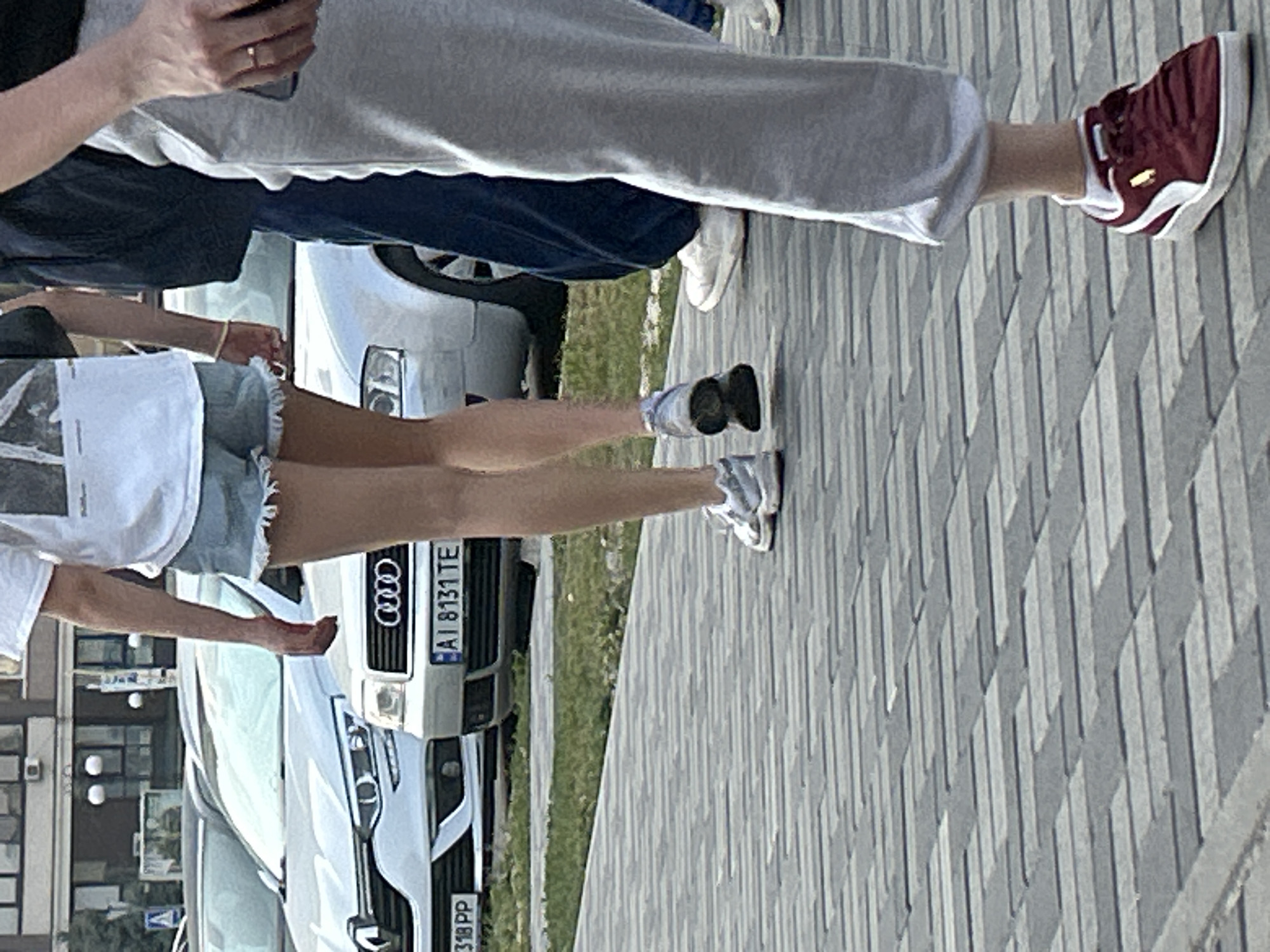The image captures a street scene in Brovary, Ukraine. In the foreground, the lower legs and feet of three individuals are visible, walking on a paved pedestrian walkway made of rectangular gray and black tiles. The person on the far right wears burgundy Puma sneakers with white accents, white socks, and light gray sweatpants. The person in the middle wears light blue denim shorts with frayed edges and light-colored athletic shoes. The third person, on the far left, is partially visible and appears to be wearing similar athletic shoes. In the background, two cars are parked. The car on the left is a silver Renault with a license plate reading "318 PP". To its right is a silver Audi with a license plate reading "AI 8131 TE". The cars are parked on a grassy area next to the walkway, with buildings visible in the distance. The overall lighting suggests it is daytime.