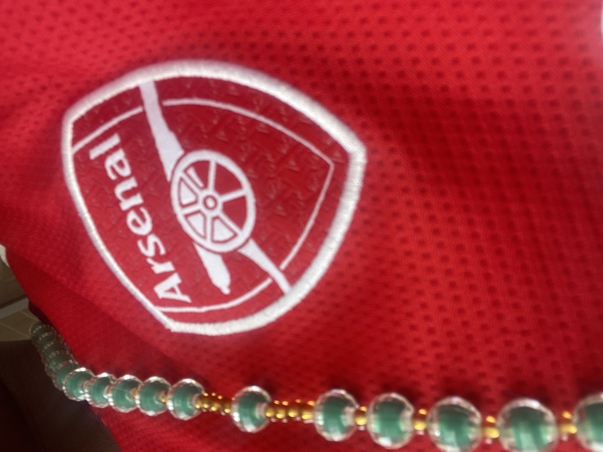 The image is a close-up shot of a red fabric with a white Arsenal Football Club crest embroidered on it. A strand of green and gold beads hangs vertically on the left side of the frame, partially obscuring the crest. The fabric appears to be a jersey, possibly sports apparel. The background is out of focus and dark, suggesting indoor lighting conditions. There are no people or any identifiable landmarks within the frame. The scene does not provide any information about the location in the city of Jos, Nigeria.