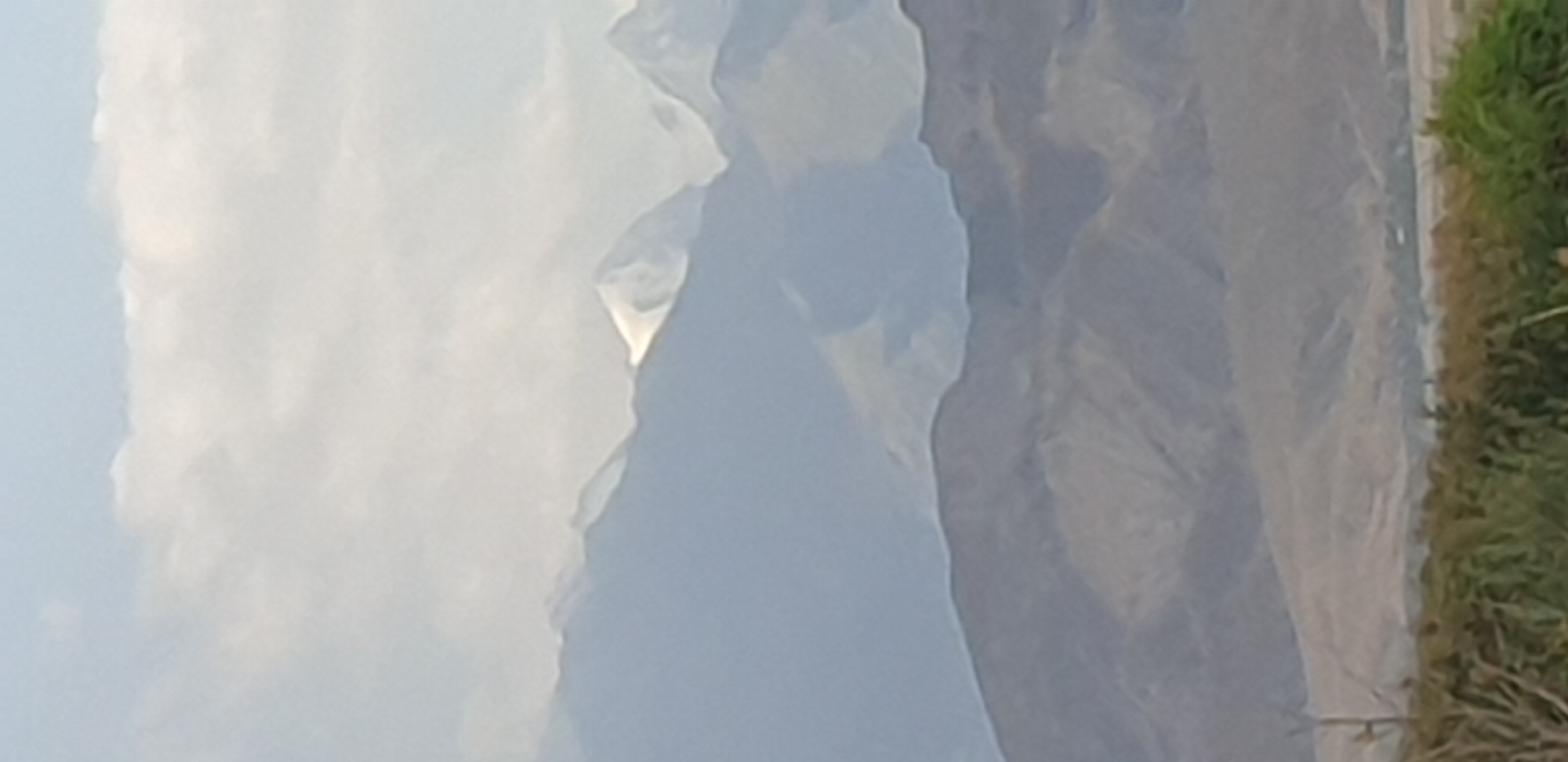 A wide-angle, outdoor, vertical shot captures a layered landscape of mountains and sky. The sky is a pale blue with soft, white clouds occupying the upper portion of the frame. Below the sky, a range of hazy, blue mountains forms the background. Further in the foreground, a second range of brown mountains is visible, with a more textured and detailed appearance. The closest layer to the viewer is a blurred strip of green foliage and dry vegetation, suggesting a foreground that is out of focus. The overall scene appears to be a natural landscape, possibly with a hint of human settlement in the distant valley between the mountain ranges. The location is identified as Leninskoye, Kyrgyzstan.
