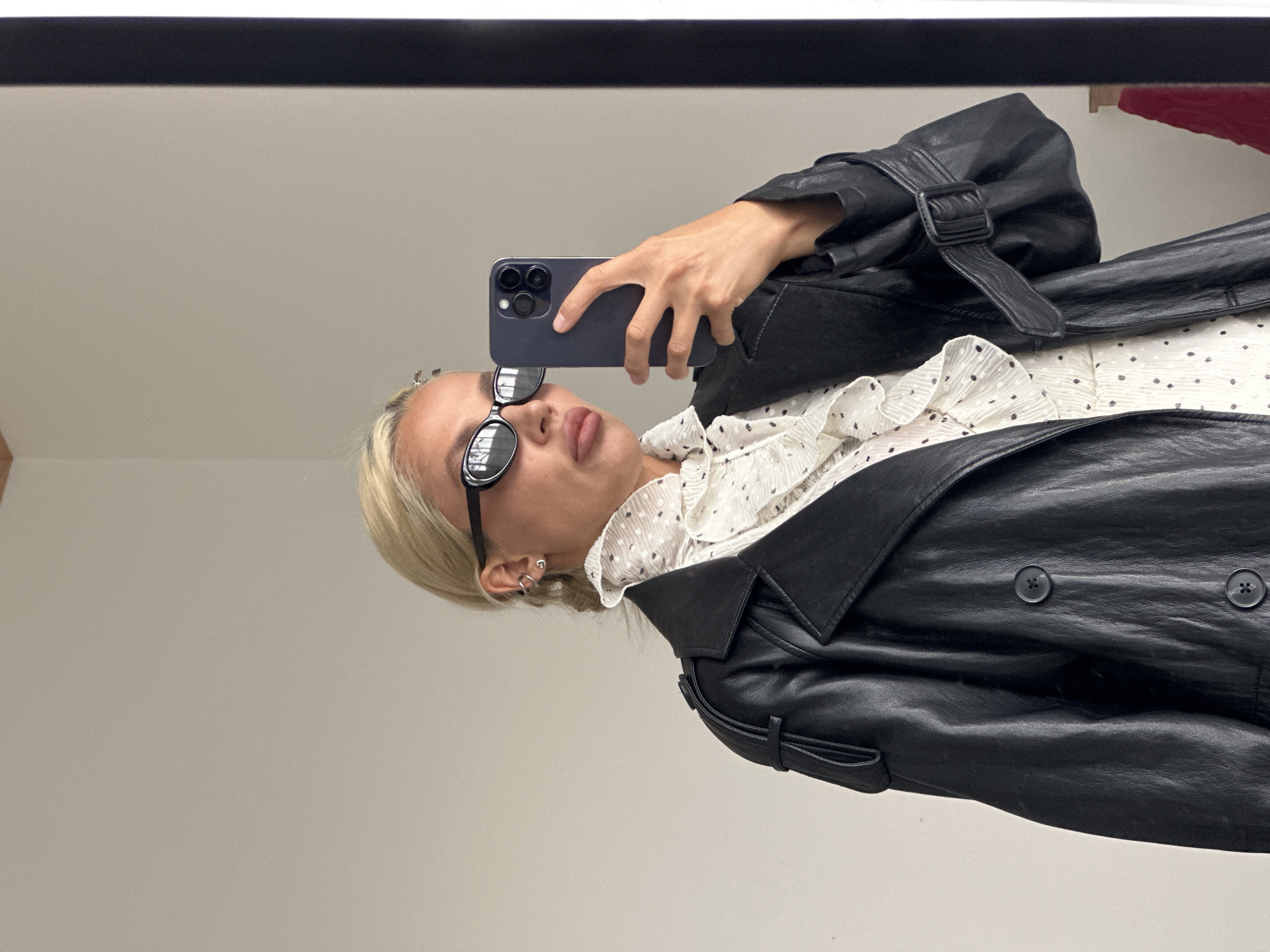 A woman with blonde hair tied in a braid and wearing sunglasses is taking a mirror selfie. She is dressed in a black leather trench coat over a white dress with black polka dots and ruffled detailing. She is holding a dark-colored smartphone in her right hand, angled to capture her reflection. The background appears to be an indoor setting with a plain, light-colored wall and a dark vertical element on the right, possibly a doorway or frame. No specific location in Kuta Selatan, Indonesia is identifiable.