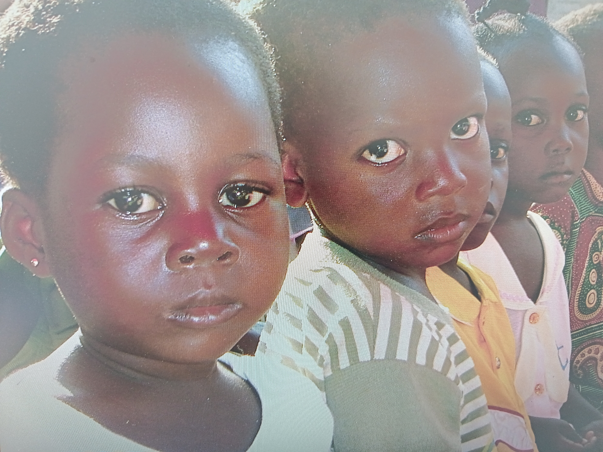 A close-up shot features a group of four young children with dark skin, potentially of African descent. The two children in the foreground are most prominent. The child on the left wears a plain white shirt and a small earring. The child on the right wears a beige shirt with white stripes. Two additional children are visible behind them. The background includes a hint of colorful patterned fabric on one of the children's clothing. The location is not identifiable as Novoselci, Bulgaria, based on the image alone.