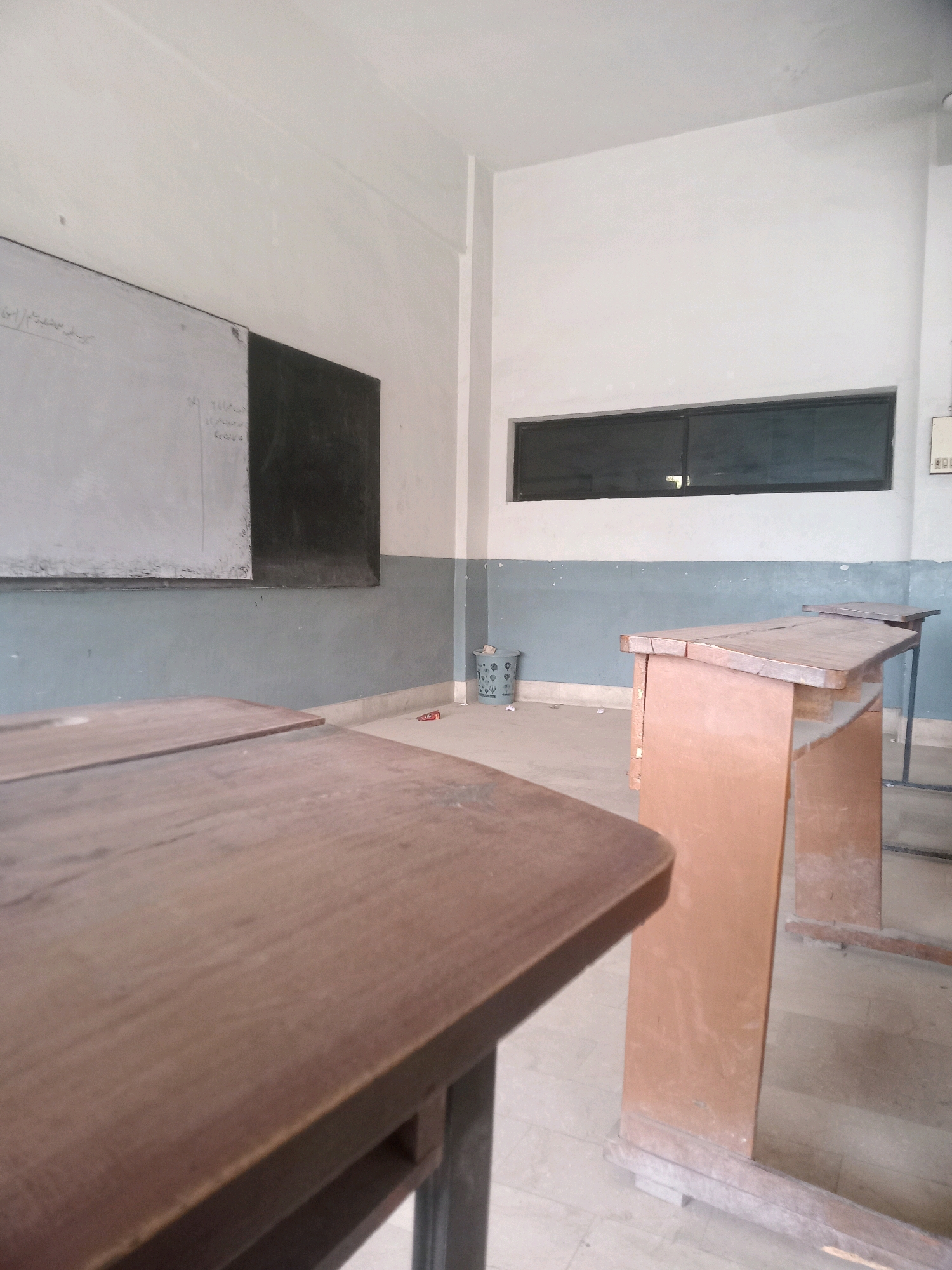 The image depicts an interior view of an empty classroom in Karachi Division, Pakistan. The room contains several wooden desks, a whiteboard with writing on it, and a window. The walls are painted white and blue, and the floor appears to be tiled. There is a waste bin in the corner and a small red object, possibly a pen, on the floor. The lighting suggests it is daytime, and the overall impression is one of a sparsely furnished and somewhat aged educational space.