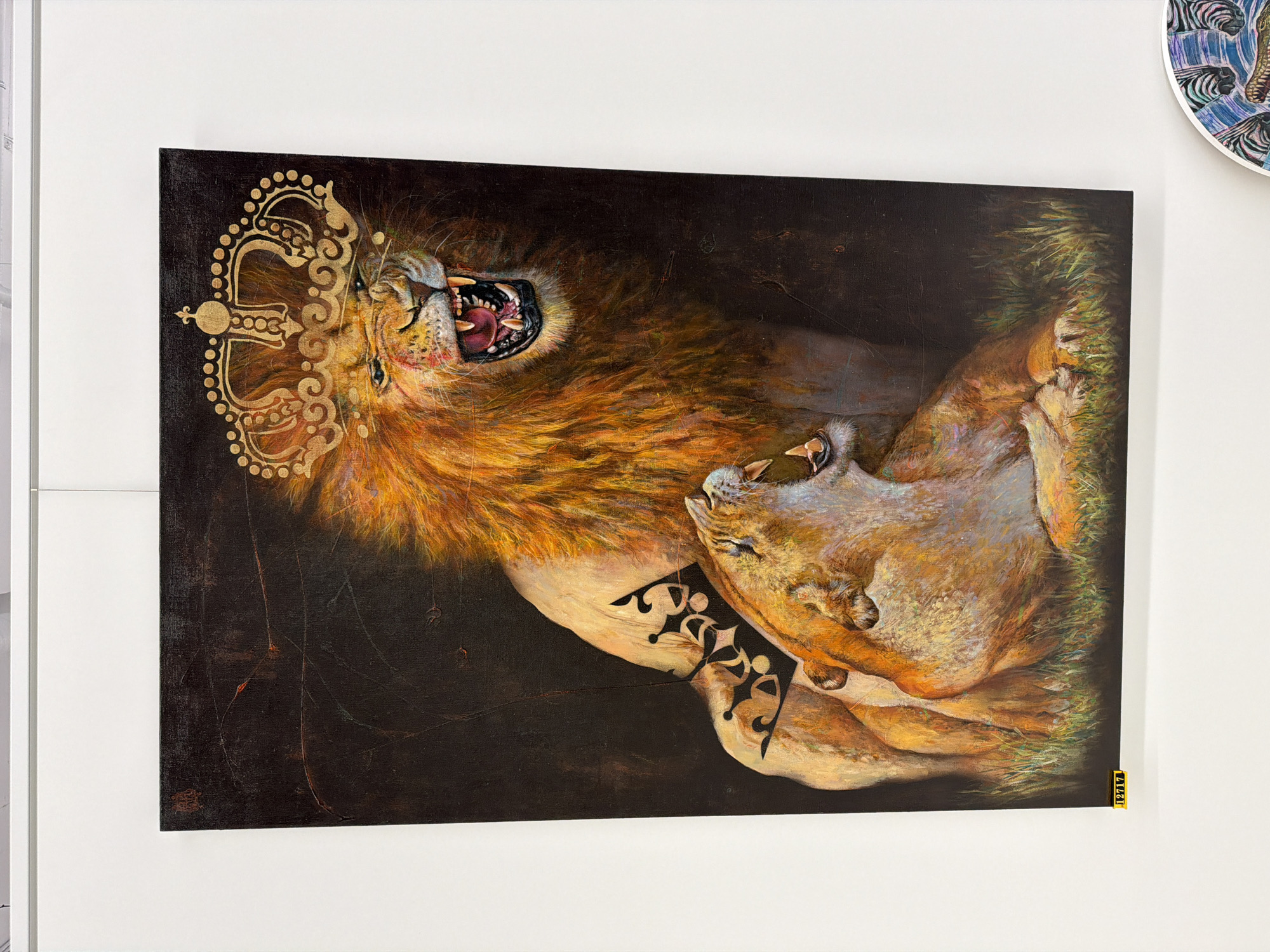 The image shows a painting of two lions, one male and one female, on display in what appears to be an art gallery. The male lion, positioned behind and slightly above the female, is depicted roaring with its mouth open, revealing sharp teeth. It wears a golden, ornate crown on its head. The female lion is lying down in front of the male, also with its mouth open, looking towards the left side of the frame. Both lions have a warm, golden-brown coloration and appear to be rendered with a textured, painterly style. The background is dark and somewhat indistinct, with hints of grass visible at the bottom. The painting is framed and hanging on a white wall. A yellow label with the numbers "12717" is attached to the bottom left corner of the painting. The painting is framed and hanging on a white wall. In the lower right corner of the image, a portion of another artwork featuring zebras and a crocodile is visible. The overall scene suggests an indoor exhibition space, likely an art gallery in Minsk, Belarus, given the context of the prompt.