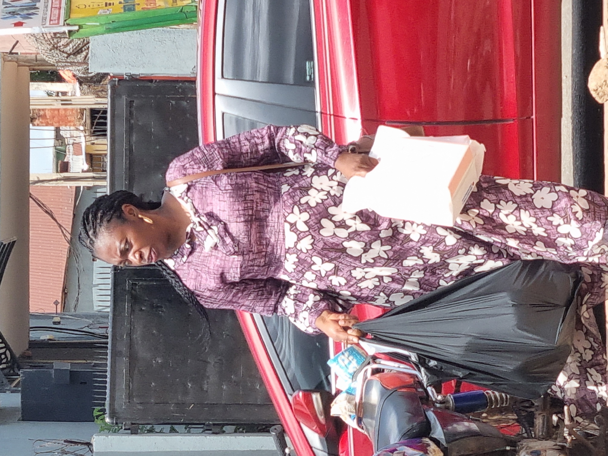 A woman is photographed in Jos, Nigeria, next to a red car and a motorcycle. She is wearing a patterned purple and white long-sleeved dress. She is holding a white cardboard box in one hand and a black plastic bag in the other. The background includes buildings, some advertisements, and a black metal structure.
