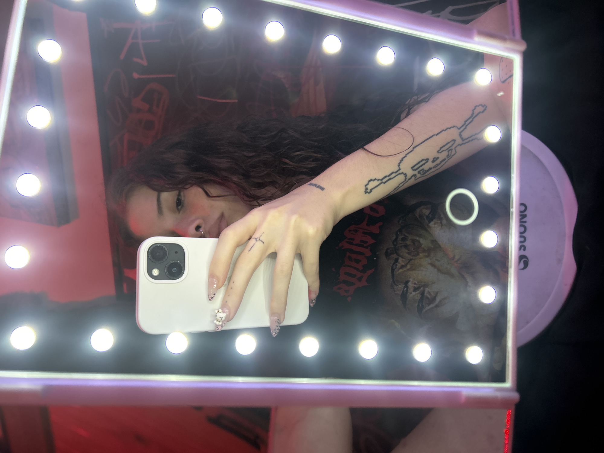A person is taking a selfie in a lighted makeup mirror, reflected in the mirror. The person has dark hair and several tattoos. A white cell phone covers part of the reflection. In the background behind the person in the mirror is a wall with graffiti. A white circular object with "SUONO" written on it is located on the right side of the reflection. It's unclear if this photo was taken in Buenos Aires, Argentina, as there are no specific landmarks or cues to confirm its location.