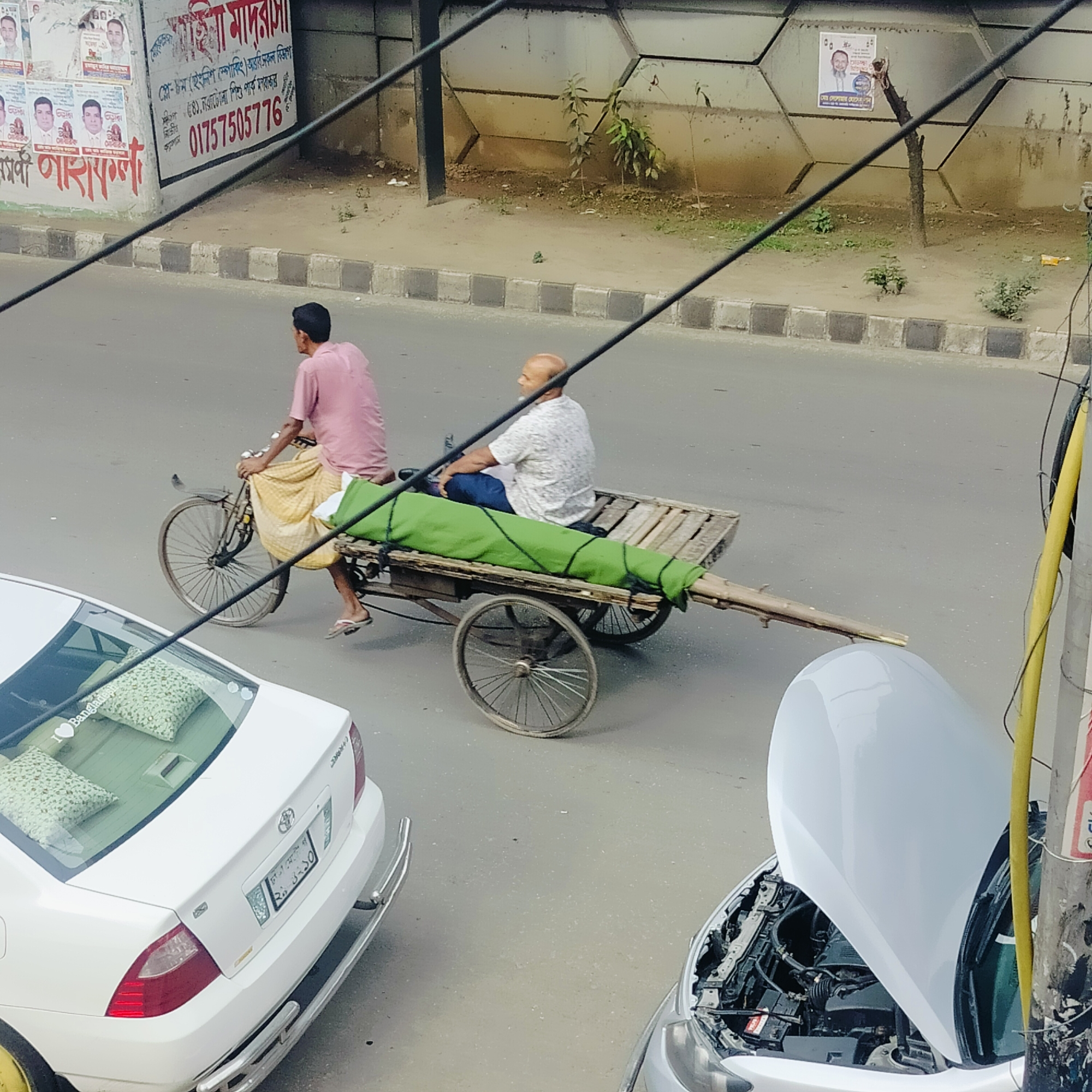 The image presents an elevated view of a street scene in Dhaka, Bangladesh. In the foreground, a white Toyota Corolla sedan is parked on the left with patterned cushions visible inside, and a light-colored car with its hood open is parked on the right. The central focus is a bicycle rickshaw with a wooden cart carrying a passenger covered in a green cloth. A man wearing a pink shirt and a yellow sarong is cycling the rickshaw, while another man in a white shirt and blue pants is sitting on the cart. The background features a wall with posters and advertisements in Bengali, including a phone number "01757505776" and the words "মাইনা মাদরাসা". Utility lines stretch across the top of the frame. The street is surrounded by sidewalks and vegetation.