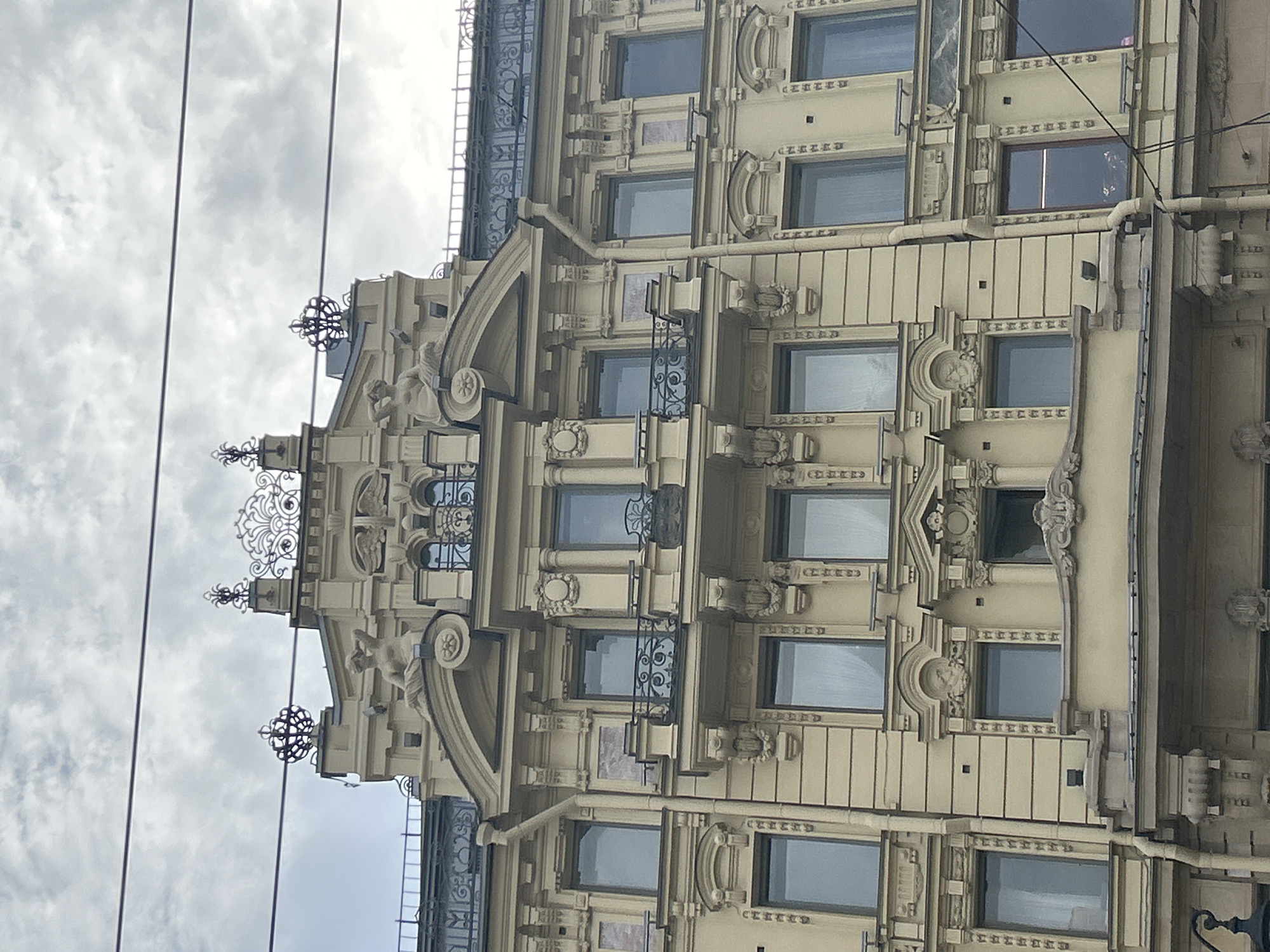 A tall, ornate, pale yellow building with multiple floors and windows dominates the frame. The architecture features neoclassical elements such as columns, pediments, and decorative sculptures. The building is topped with elaborate ironwork and sculptures of winged figures and crowns. Several power lines are visible against a cloudy, grey sky. The image appears to be a photograph of the facade of a building in an urban setting, possibly a historic district.