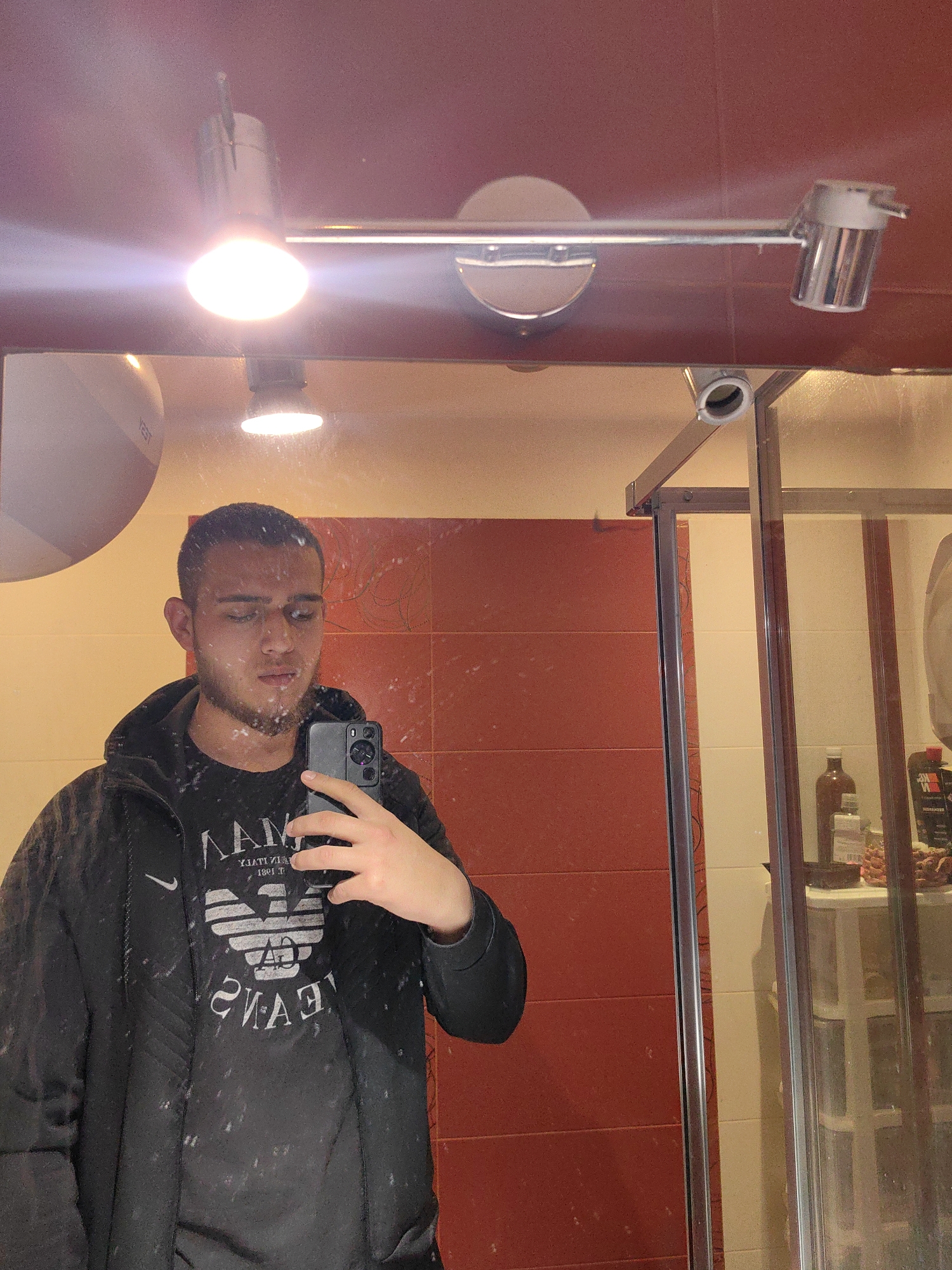 A man is taking a selfie in a bathroom mirror. He is wearing a black jacket with an Armani Jeans shirt underneath. The bathroom has a red tiled wall, a shower cubicle with toiletries on a shelf, and a light fixture with three spotlights. The exact location in Sofia, Bulgaria cannot be determined from the image.