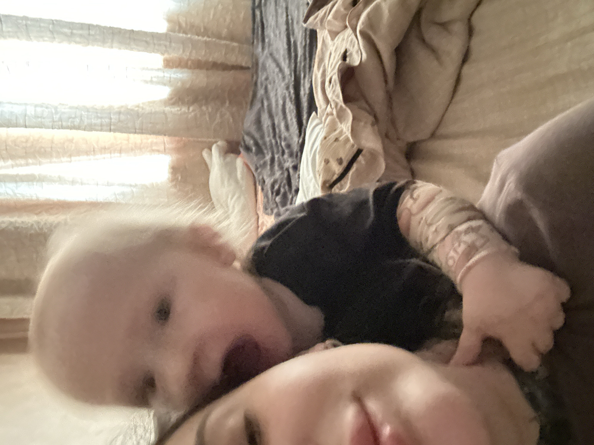 An indoor, close-up, self-portrait-style photograph shows a woman and a baby. The woman is in the foreground on the left, with her face mostly visible, looking towards the camera. The baby is behind her, to the right, with its head tilted, appearing to be interacting with the woman. The baby's mouth is open. Both subjects are in focus, though the baby is slightly more blurred. The background consists of a bed with blankets and curtains visible near a window, suggesting a domestic setting. The lighting appears to be natural light coming from the window. There are no discernible landmarks or elements that would indicate the location within the city of Kyiv, Ukraine.