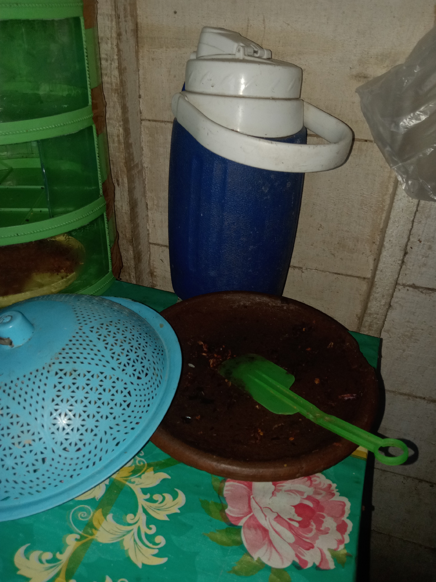 This is a still life photograph taken indoors, likely in a kitchen or storage area, with a focus on several household items. The scene is illuminated by artificial light, casting shadows.
In the center of the frame stands a blue and white insulated water jug with a white handle. To its left is a light blue colander with a perforated design. In front of the jug and slightly to the right is a brown, shallow mortar containing some dark residue and a green plastic spatula resting across it. The items are placed on a table covered with a teal-colored tablecloth featuring a floral pattern with large pink and white flowers and green leaves.
To the far left of the image, a portion of a green plastic shelf unit with multiple tiers is visible. The background consists of a pale, textured wall, possibly made of wood or concrete. A crumpled plastic bag is present in the upper right corner of the image.
The photograph does not contain any people or depict any actions or interactions. The environment appears to be a residential setting, possibly in Stiaji, Indonesia, given the context provided. The overall impression is one of everyday domestic objects arranged in a functional space.
