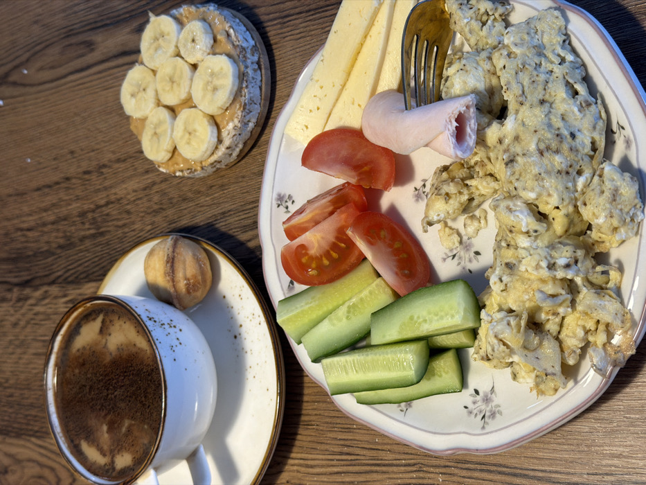 This image displays a breakfast or brunch meal set on a dark brown wooden table, originating from Perm, Russia. In the upper left, a white ceramic cup with a brown rim and speckles, containing dark coffee with light foam, rests on a matching saucer. Adjacent to the cup, a single light brown, shell-shaped pastry, likely an "Oreshki," is present. To the upper right, a round rice cake is topped with a layer of peanut butter and nine slices of banana, sitting on a small wooden coaster. In the foreground, a white ceramic plate with a delicate floral pattern and a scalloped pink-brown edge holds multiple food items. These include approximately six green cucumber sticks, five red tomato wedges, several rectangular slices of yellow cheese, and a substantial portion of scrambled eggs seasoned with dark flecks. Two light pink rolled slices of processed meat, identified as ham, are positioned on the plate, with one piece pierced by a silver fork exhibiting brown-toned tines. No individuals are visible, and no active interactions are depicted; the scene shows a static arrangement of food prepared for consumption.