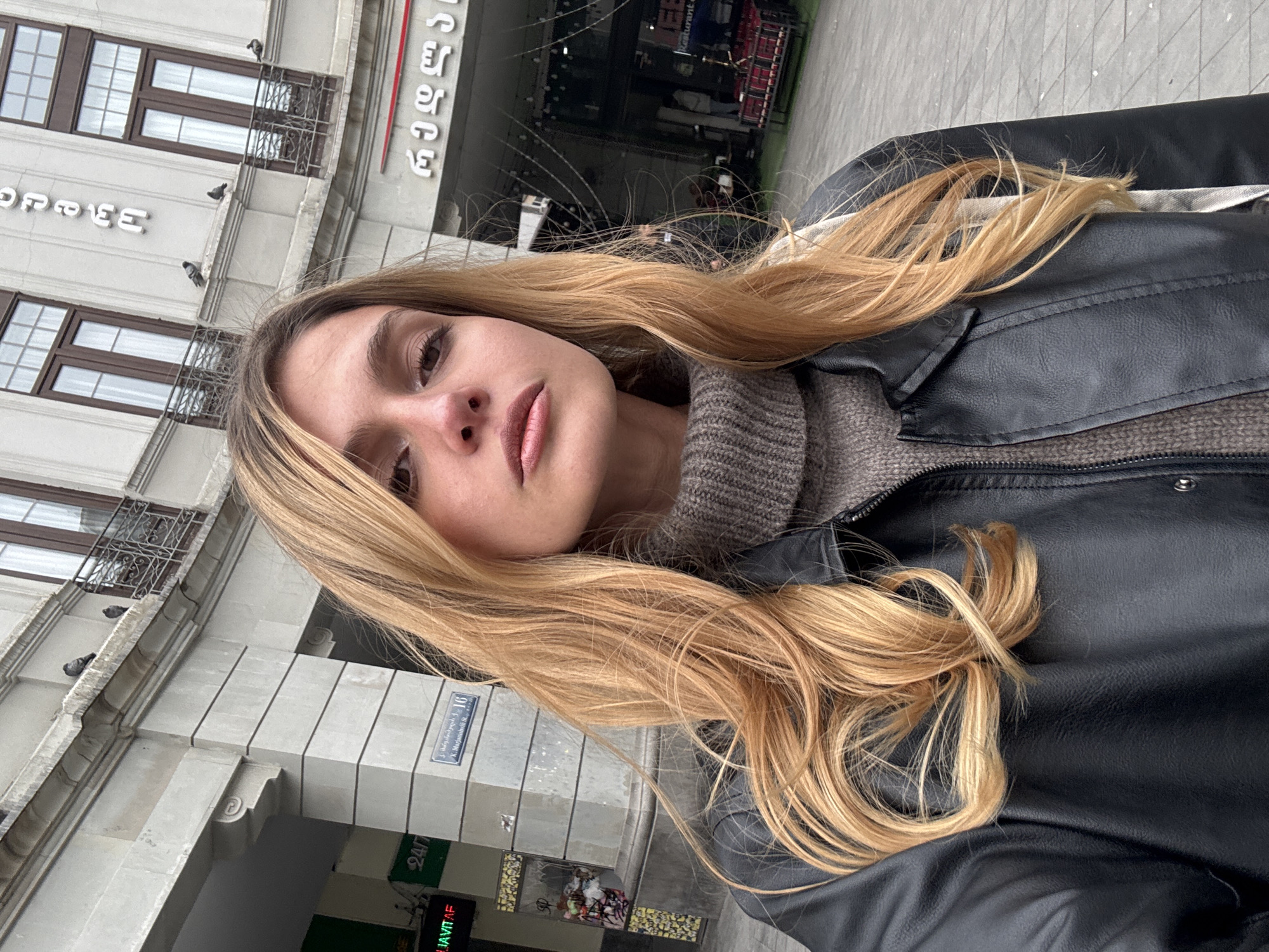 A young woman with long, flowing blonde hair is captured in a tilted selfie. She is wearing a black faux-leather jacket over a grey, ribbed turtleneck sweater. Her gaze is directed towards the camera, with a neutral expression and lips slightly parted, accented by light pink lipstick.
The background reveals an urban setting in Tbilisi, Georgia. The architecture consists of light-colored, multi-story buildings featuring dark-framed windows and decorative iron balconies. Several pigeons are visible perching on the ledges of one building. An arched entryway is also part of the street-level view.
Notable details confirming the location include Georgian script on various signs. Text visible on buildings includes "კომფორტი" (Komforti), and another sign displaying "24/7" along with "ჯავითა" (Javita). Most definitively, a blue street sign reads "კ. მარჯანიშვილის ქ. 16," which translates to "K. Marjanishvili Street 16," explicitly identifying the specific street. The lighting indicates it is daytime, likely overcast, providing soft illumination to the scene. The image captures a casual moment in a vibrant city environment.