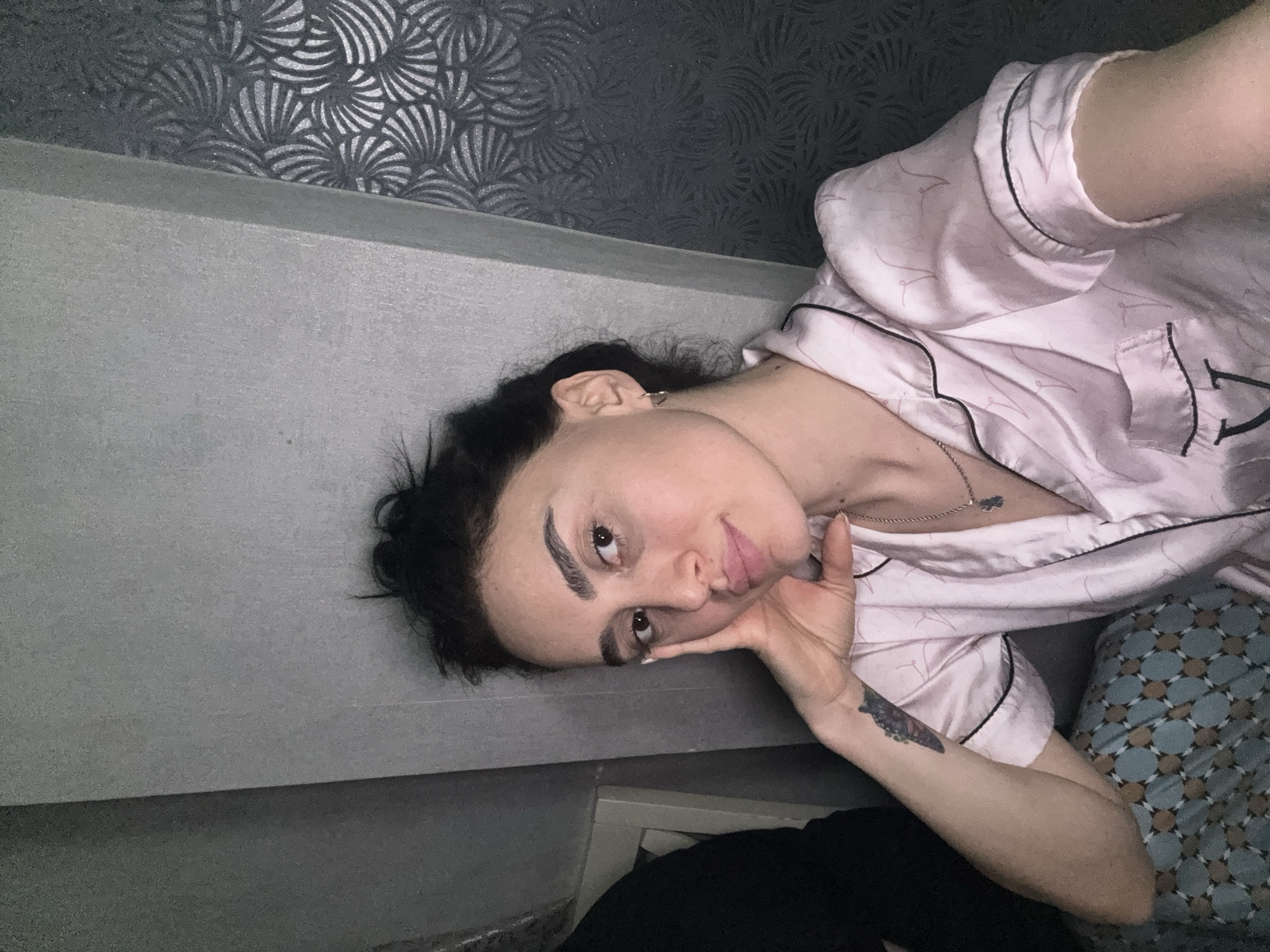 A young woman is lying on a gray surface, possibly a bed or bench, in an indoor setting. She is wearing light pink pajamas with black trim. She has dark hair pulled back and is resting her face on her hand. A tattoo is visible on her forearm. The background includes a wall with a textured silver-and-dark-gray pattern and a section of a blue-patterned pillow or blanket. The location is not identifiable as Tbilisi, Georgia based on the provided image alone.