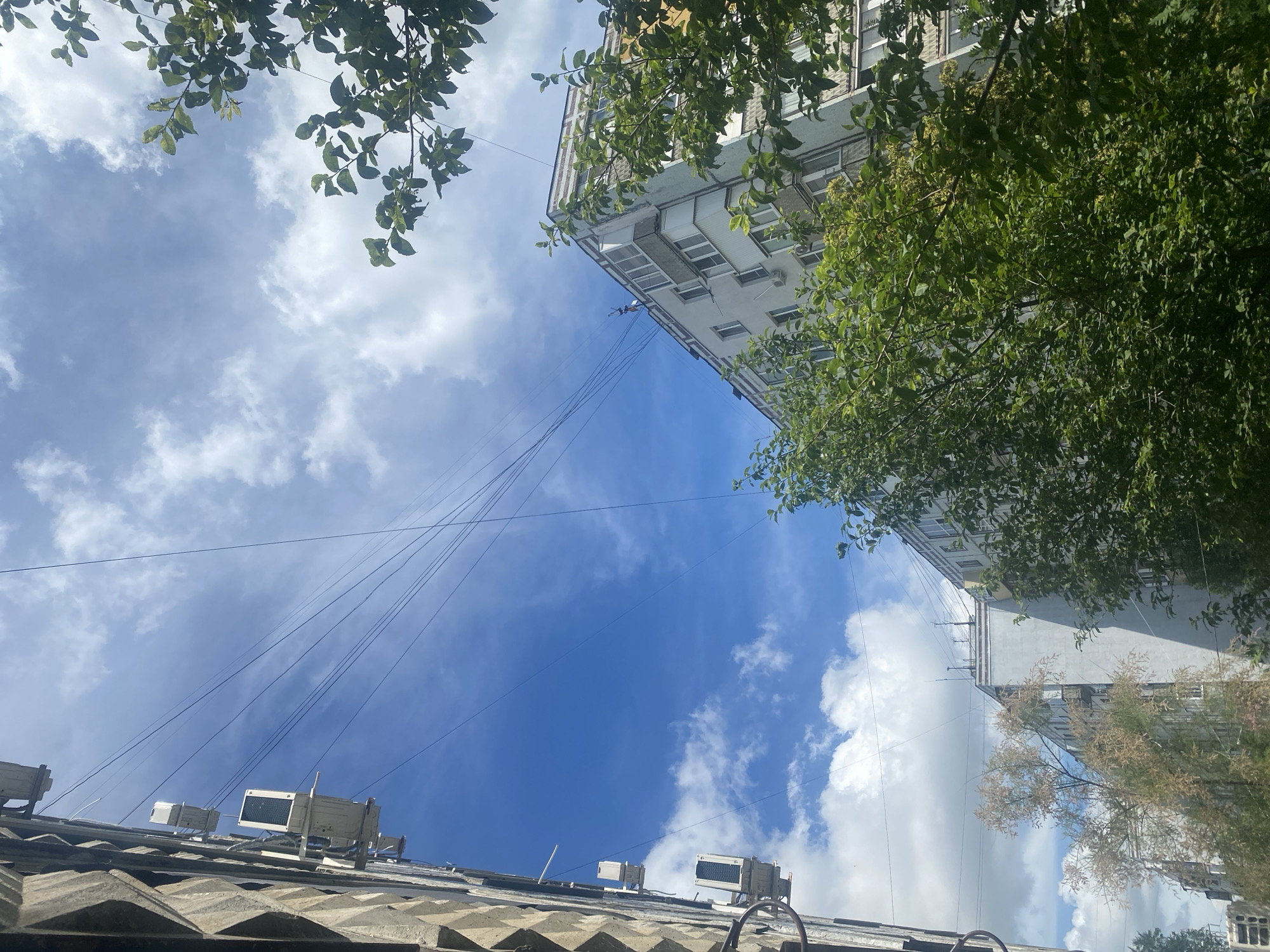 The image captures a view of an urban residential area, likely in Samara, Russia, looking upwards towards the sky. The scene is dominated by a bright blue sky with scattered white clouds. On the left side of the frame, a textured concrete building facade is visible, with several outdoor air conditioning units mounted on its exterior. Overhead, a complex network of thin wires crisscrosses the sky. To the right, another multi-story apartment building with a light-colored facade and balconies can be seen, partially obscured by lush green trees. The trees are full of leaves, suggesting a warmer season. The overall impression is one of a typical city dwelling environment during daylight hours. There are no people visible in the image.