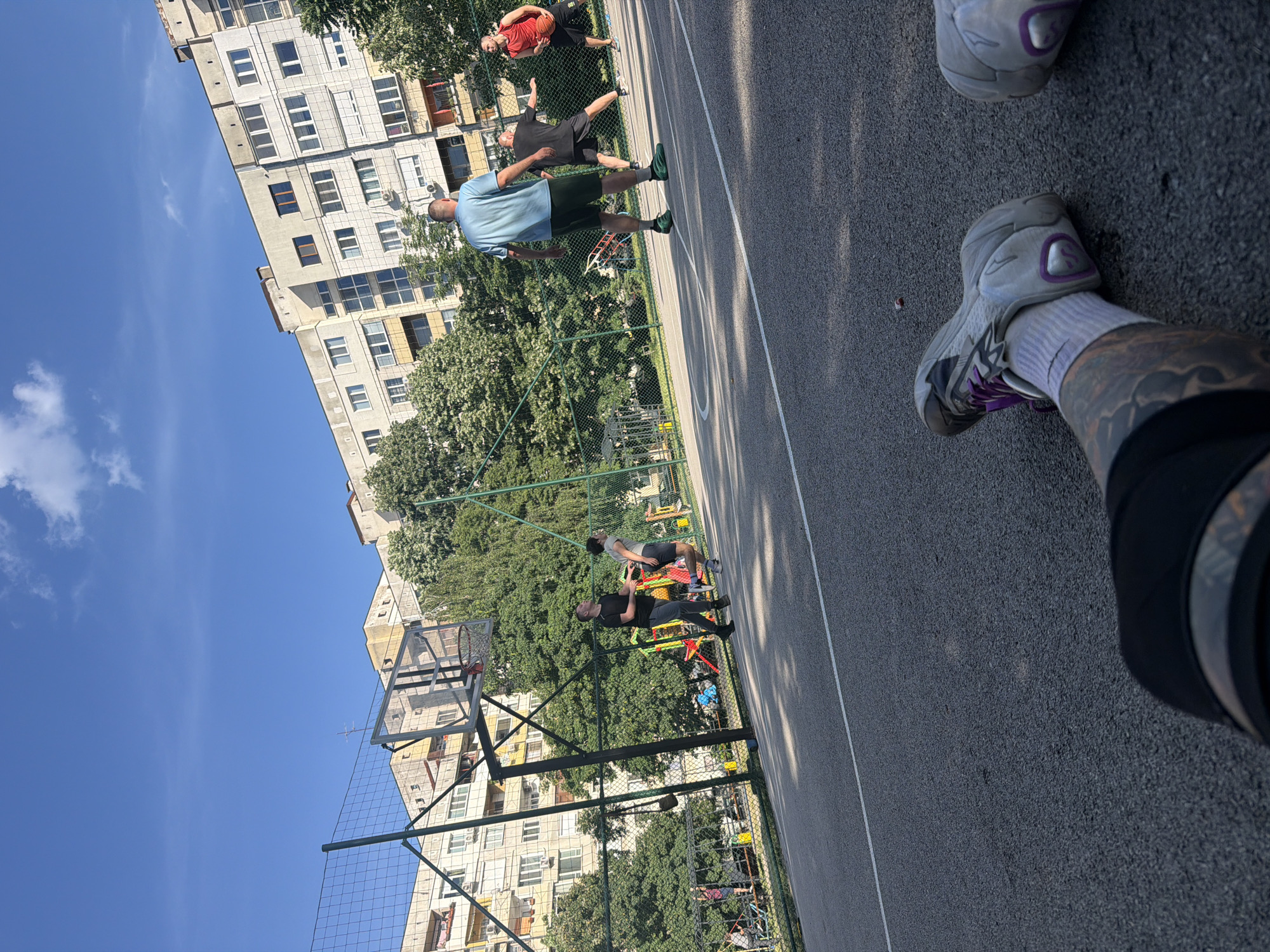 A street-level, low-angle shot captures a basketball court adjacent to a high-rise apartment building, under a blue sky in Sofia, Bulgaria. The photographer's legs and feet are visible in the foreground, clad in white socks and white and grey sneakers with purple accents. Three individuals are spaced across the court. One is in a light blue shirt and dark shorts, and another is in a red shirt and dark shorts, while another individual is in black athletic clothing. A basketball hoop and net stand on the left side of the frame. The basketball court is surrounded by a green chain-link fence. Trees fill the backdrop, interspersed with buildings that appear to be part of an urban residential area.