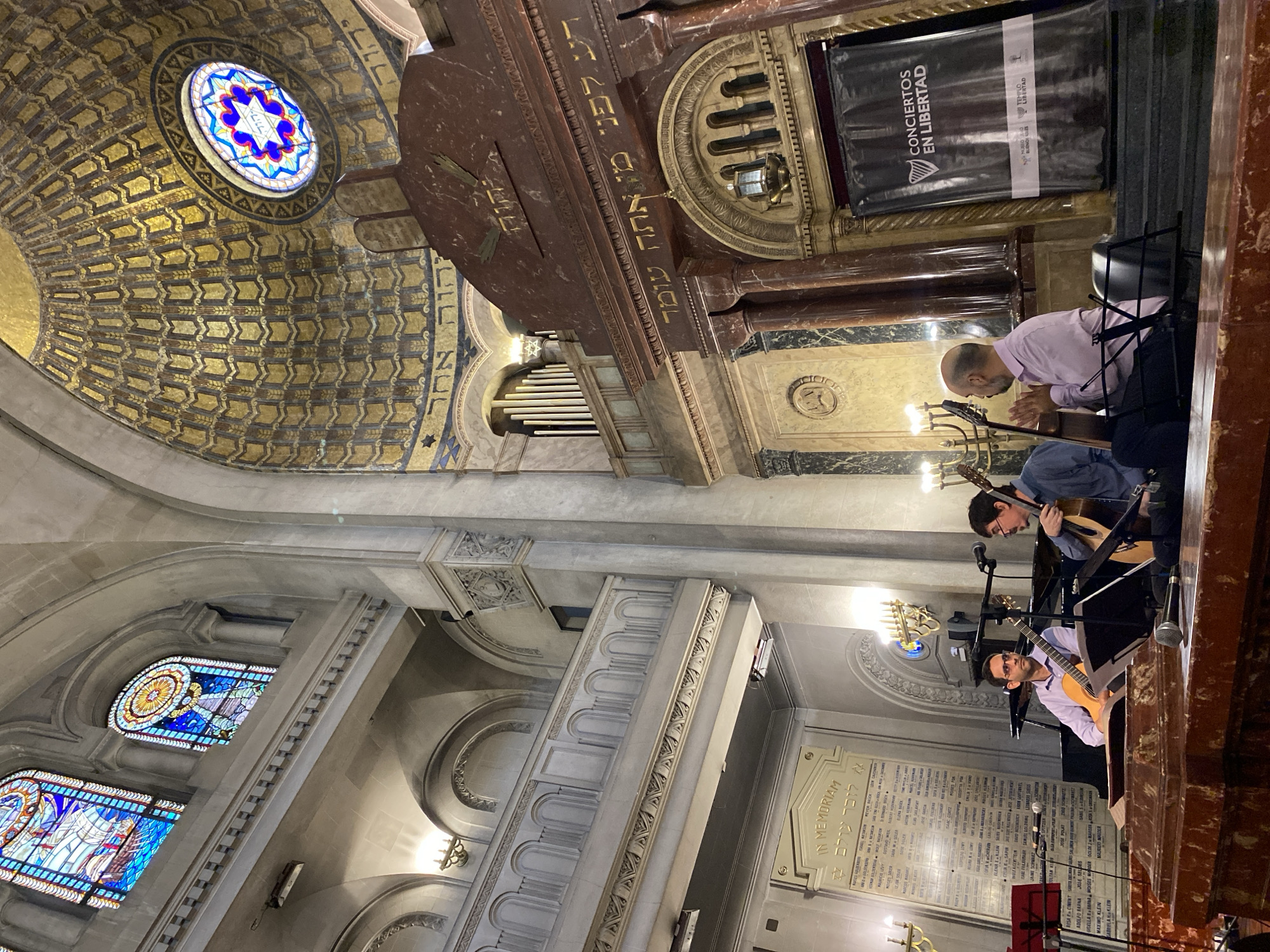 The image shows the interior of a building, specifically Templo de la Congregación Israelita Argentina (a synagogue) located in Buenos Aires, Argentina. The architectural details, including the dome with a stained glass window featuring a Star of David, the ornate walls, and the Hebrew inscriptions above the platform confirm this. A banner titled "CONCIERTOS EN LIBERTAD" ("Concerts in Freedom") is visible. Two men are seated on a platform, playing classical guitars. Another man in a pink shirt is seated behind them, presumably managing the event. An "IN MEMORIAM" plaque with names listed on it is placed on the wall.