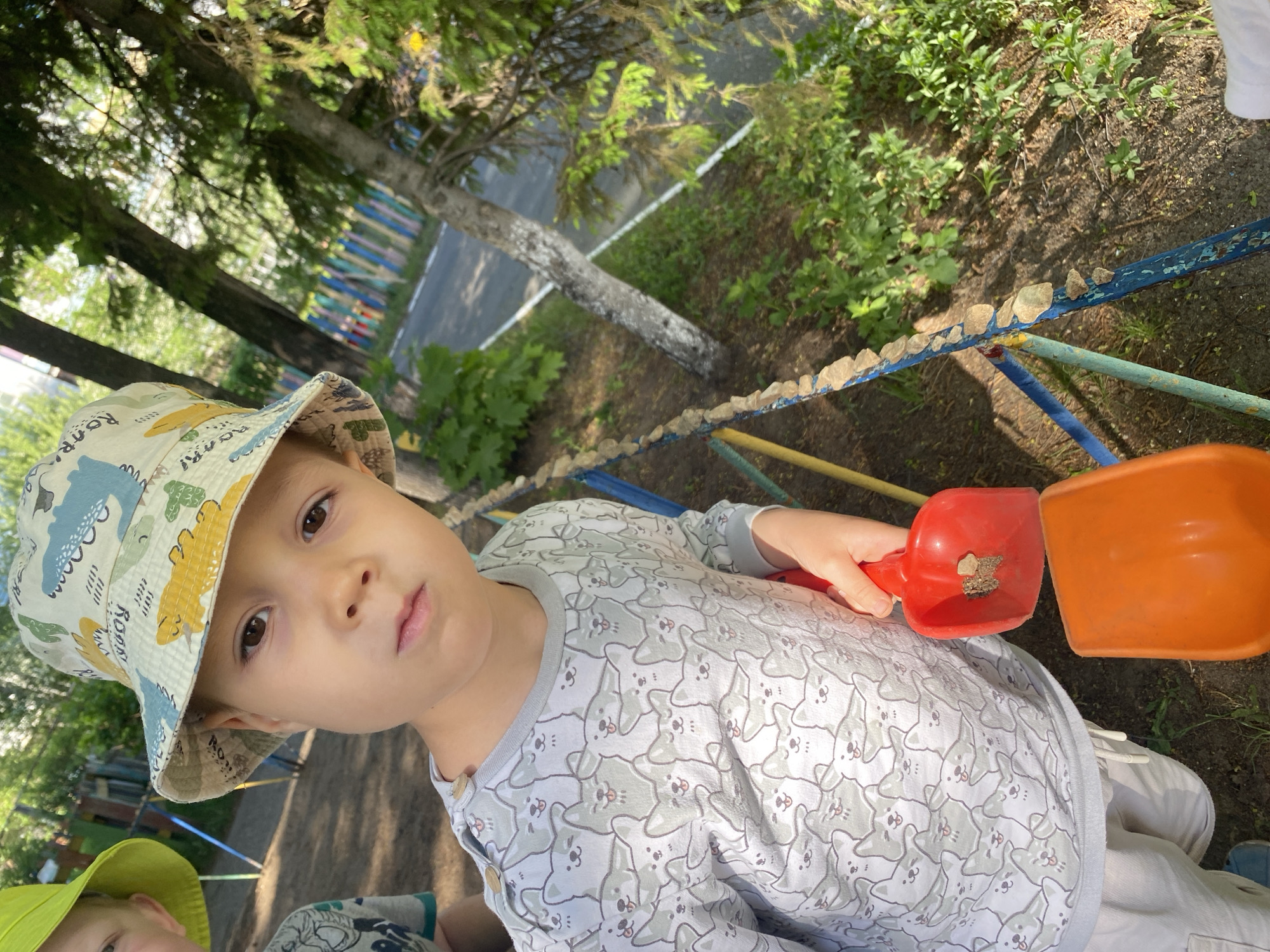 A young child, likely a boy, is the primary subject. He wears a light-colored hat patterned with dinosaurs and a long-sleeved shirt with a repeating dog-like pattern. He holds a small red shovel containing what appears to be a piece of rock or debris. An orange shovel is positioned next to it.
The setting appears to be outdoors, likely a playground or park. There is a metal fence with rocks placed on top of it. Trees and foliage are visible in the background.
Based on the geotags, this image was taken in the city of Chaikovsky, Perm Krai, Russia.