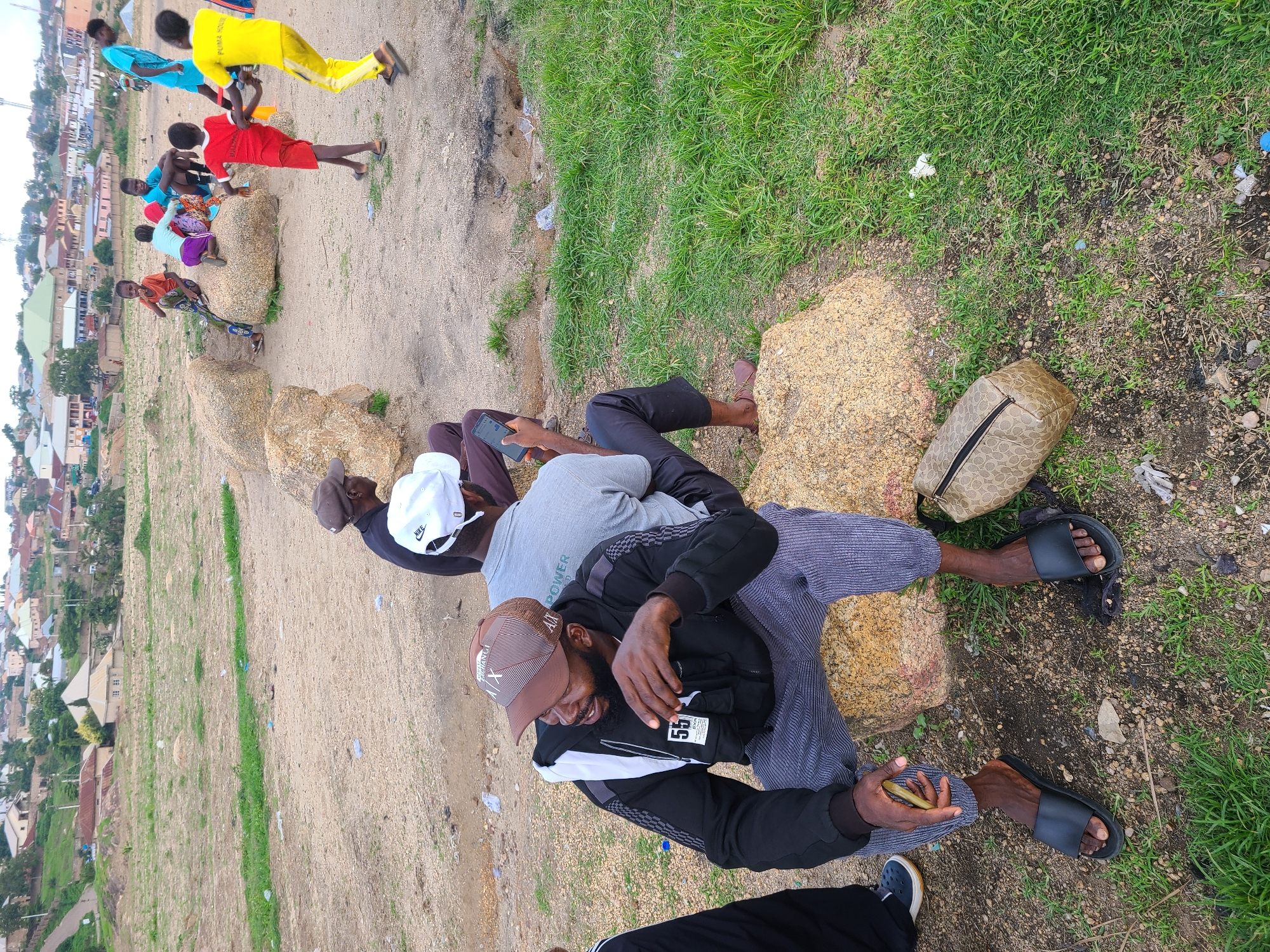 In Jos, Nigeria, a group of young men and boys are gathered outdoors on a slightly elevated, dry, and sandy area with patches of grass. The scene appears to be a casual get-together, possibly after a game or activity, as some are dressed in athletic wear.
In the foreground, three men are seated on or near large rocks. One man, wearing a black jacket with grey accents and a brown baseball cap, is looking down and smiling while holding a mobile phone. Another man, in a grey t-shirt and black pants, is leaning back, also holding a phone. A third man, in black pants and black sandals, is seated next to him. All three are focused on their phones or engaged in conversation.
In the background, a group of boys in brightly colored clothing are interacting. One boy wears a yellow shirt and pants, another a red shirt, and another a blue shirt. They appear to be playing or resting near more large rocks and a patch of grass.
The setting is characterized by its dry, dusty ground, scattered rocks, and areas of green grass. In the distance, a densely populated urban or semi-urban area with numerous buildings is visible, suggesting the location is on the outskirts of a town or city. The lighting and soft shadows indicate it is daytime, likely late afternoon or early evening. The atmosphere seems relaxed and communal, with individuals absorbed in their devices or socializing. No specific text is clearly legible beyond a tag on the jacket of the man in the foreground that appears to say "55".