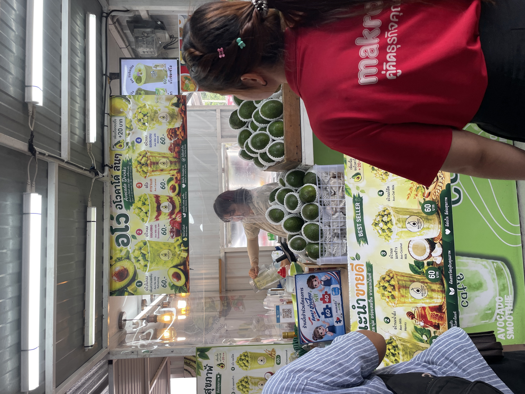 The image captures an outdoor beverage stall in Pattaya City, Thailand, specializing in avocado drinks. In the foreground, the backs of two people are visible. One person, wearing a blue and white striped shirt, stands on the left, partially obscuring a sign. The other person, wearing a red t-shirt with "makro" and Thai text printed on the back, is in the right foreground, looking towards the stall.
The stall itself is characterized by a clear plastic sheeting enclosure, likely for hygiene or weather protection, behind which a woman in a light-colored shirt is actively preparing a drink, pouring liquid from a blender jug into a cup. A display board behind her showcases various avocado drinks with Thai and English descriptions and prices. The drinks include options like "Avo-Milk," "Avo-Milk with Almond," "Avo Caramel," "Avo-Coconut Milk," and "Avo-Oat Milk," with prices listed as 60 Baht. Fresh avocados are prominently displayed in wooden crates and baskets, emphasizing the fresh ingredients. Additionally, there are smaller promotional signs, including one with cartoon characters and text related to a business partnership or promotion. The overall environment suggests a bustling, informal street food or market setting.