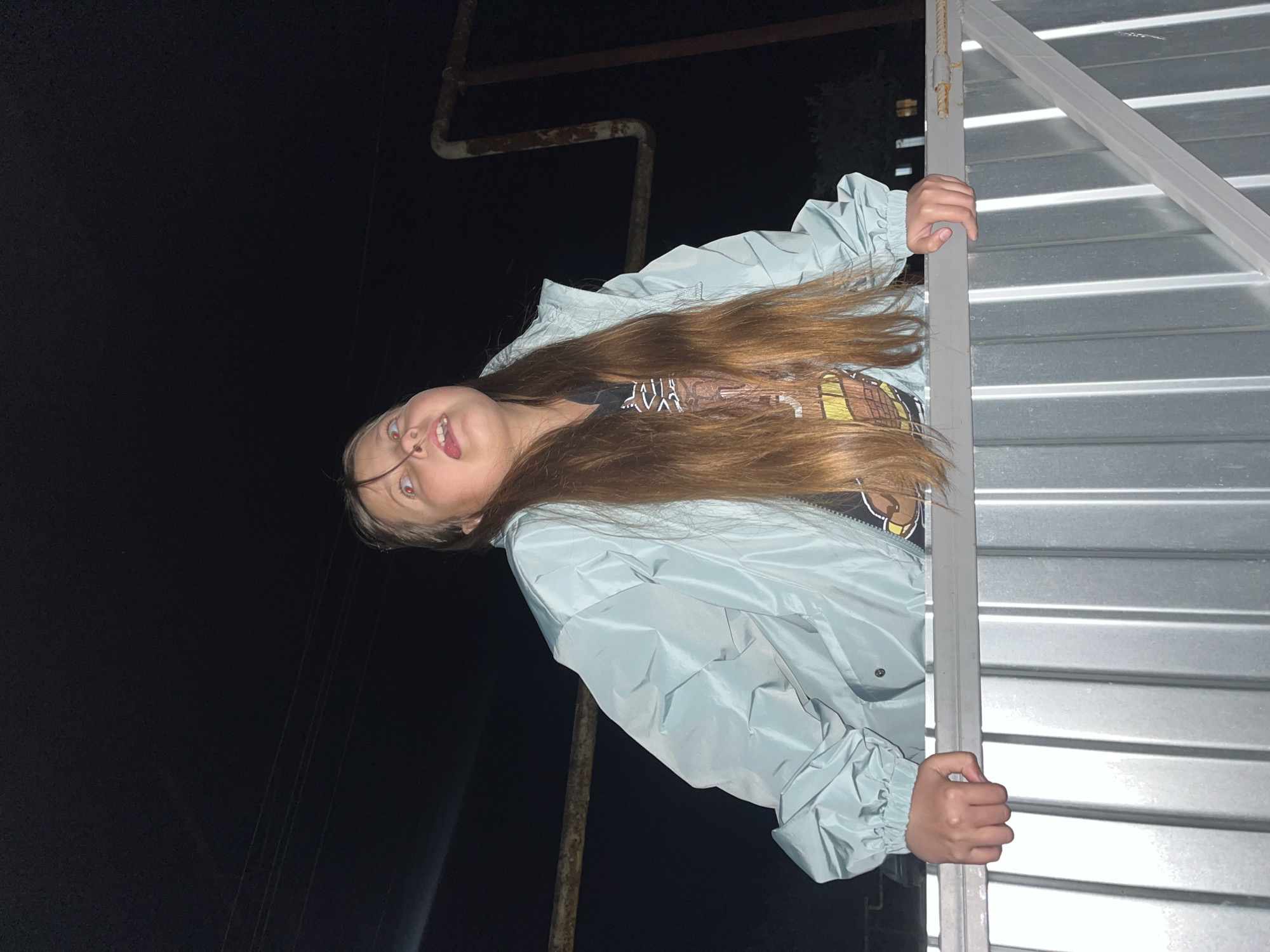 A young person is outside at night, possibly in Novopokrovskaya, Russia, given the location request. They are wearing a light blue jacket and holding onto a metal structure resembling a fence or gate. The person has long brown hair and is sticking their tongue out. Overhead, there is a pipe and what appears to be power lines running into the dark sky.